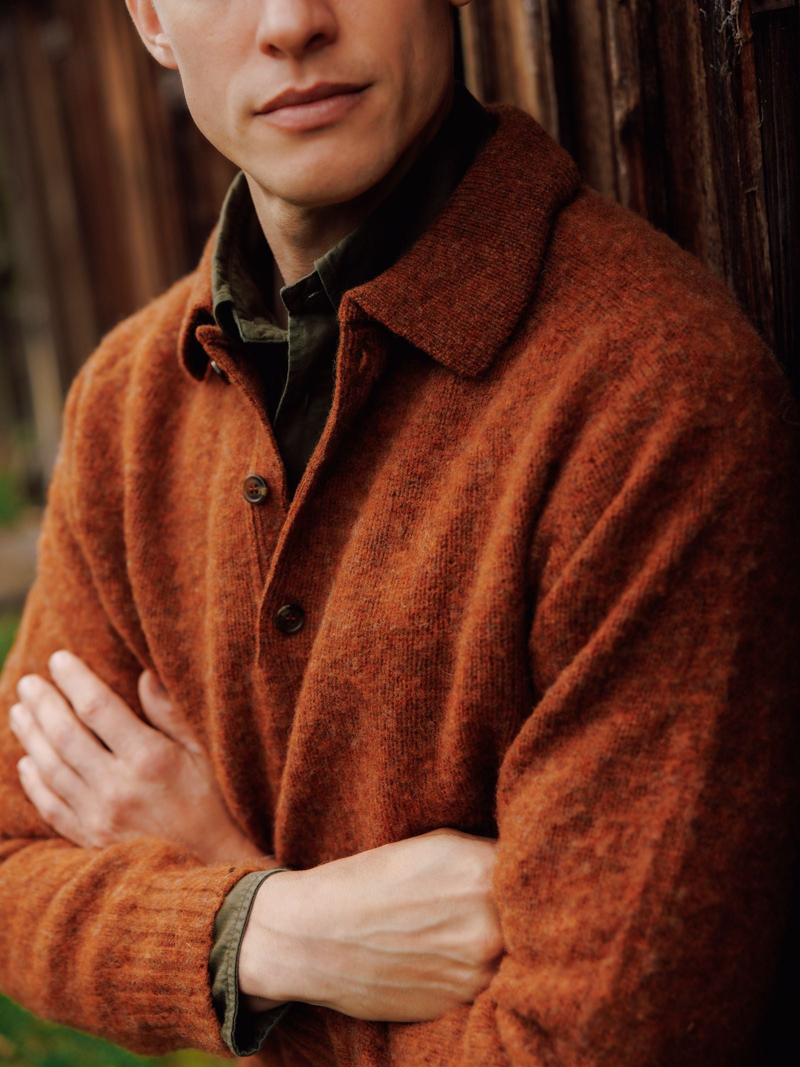 A person with light skin wears a rust Brushed Shetland Rugby Jumper by Campbell's of Beauly over a green collared shirt, standing arms crossed before a wooden wall. Their face is partly visible, looking slightly aside.