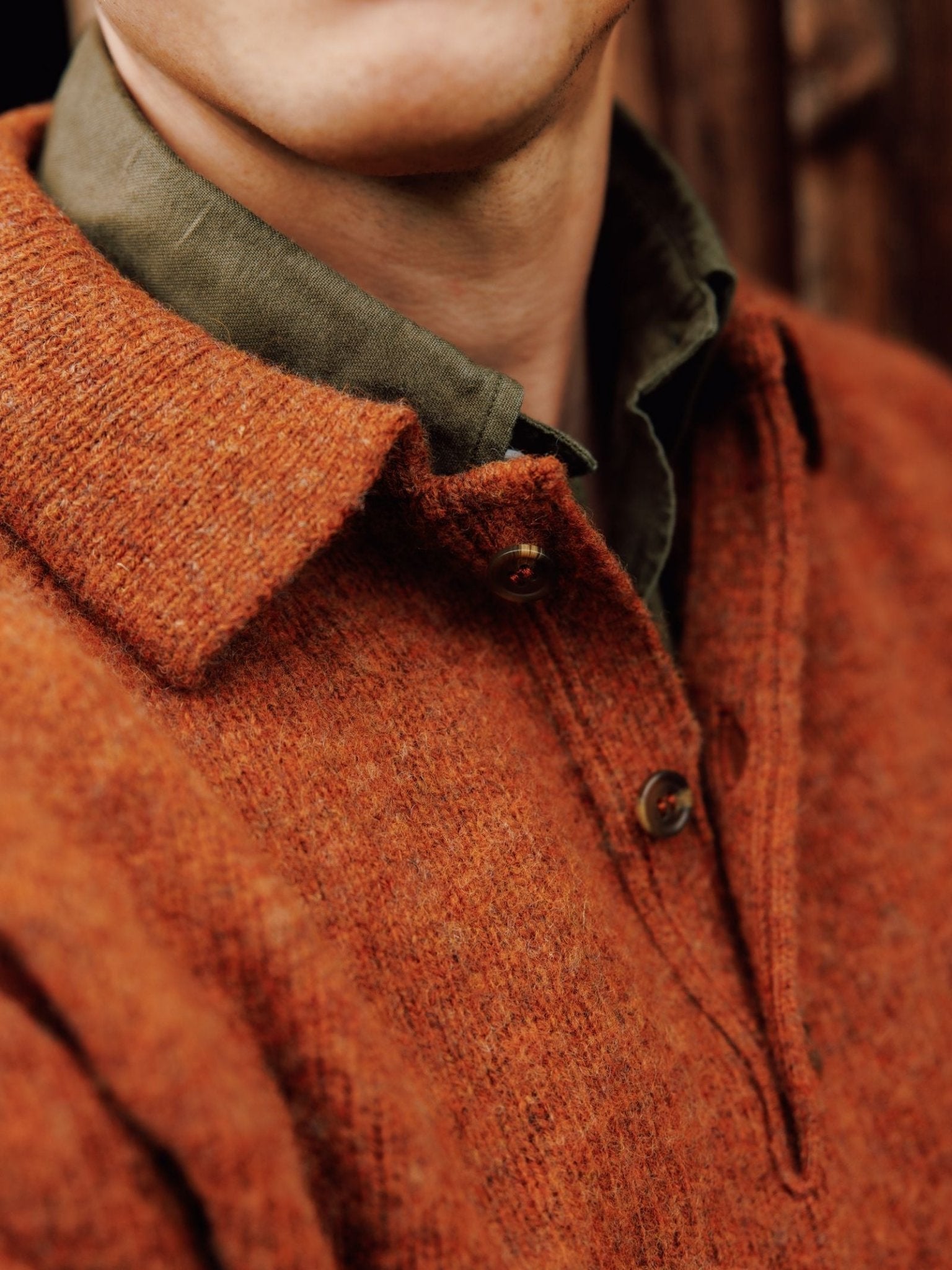 Close-up of a person in an orange Campbell's of Beauly Brushed Shetland Rugby Jumper over a green collared shirt. The image focuses on the jumper’s buttons and collar, with the lower part of the wearer’s face partly visible.
