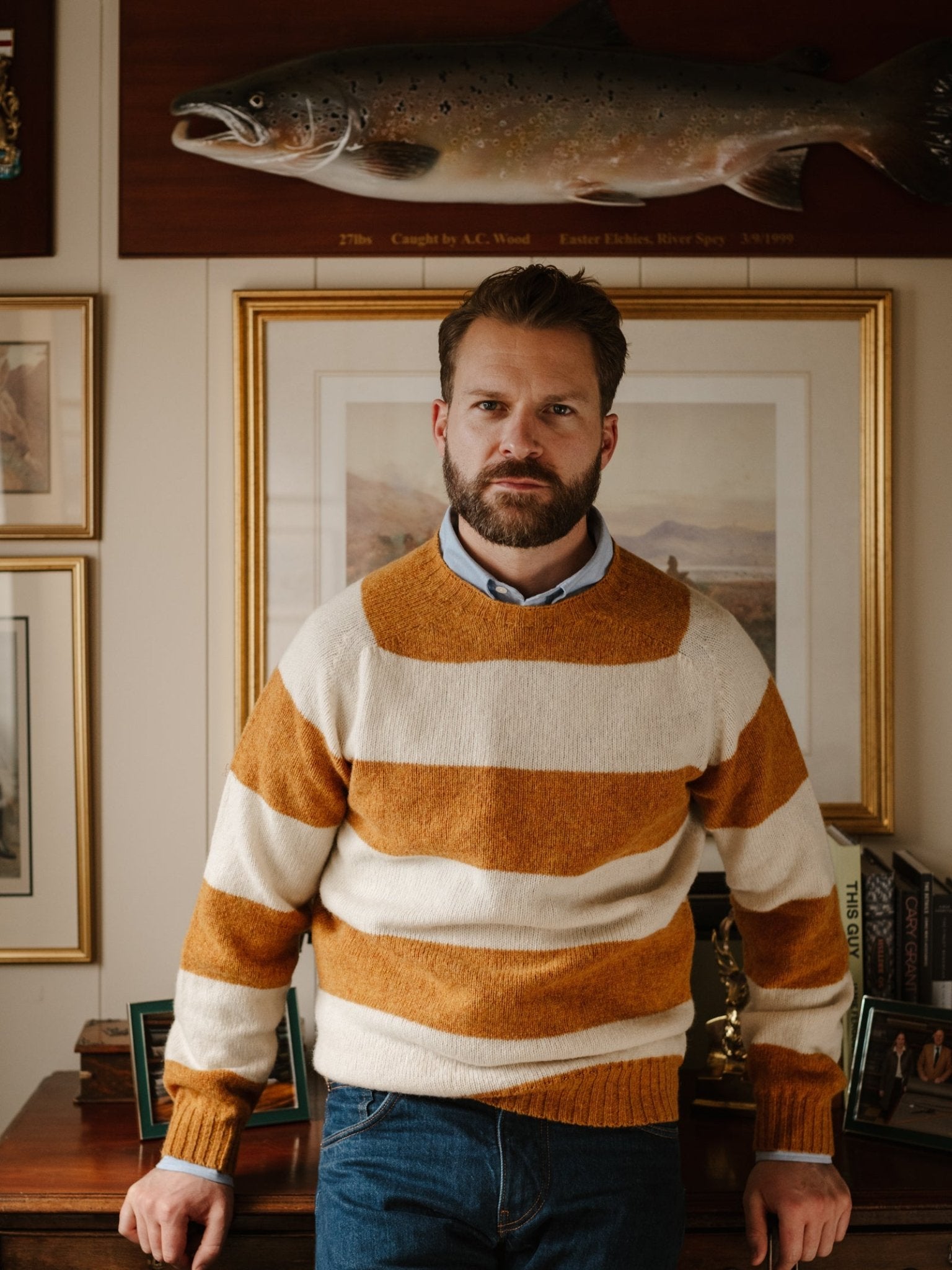 A bearded man indoors leans against a desk, surrounded by framed pictures and a mounted fish, wearing the Shetland Stripe Crew Jumper by Campbell's of Beauly—a nod to his classic taste in Scottish knitwear.