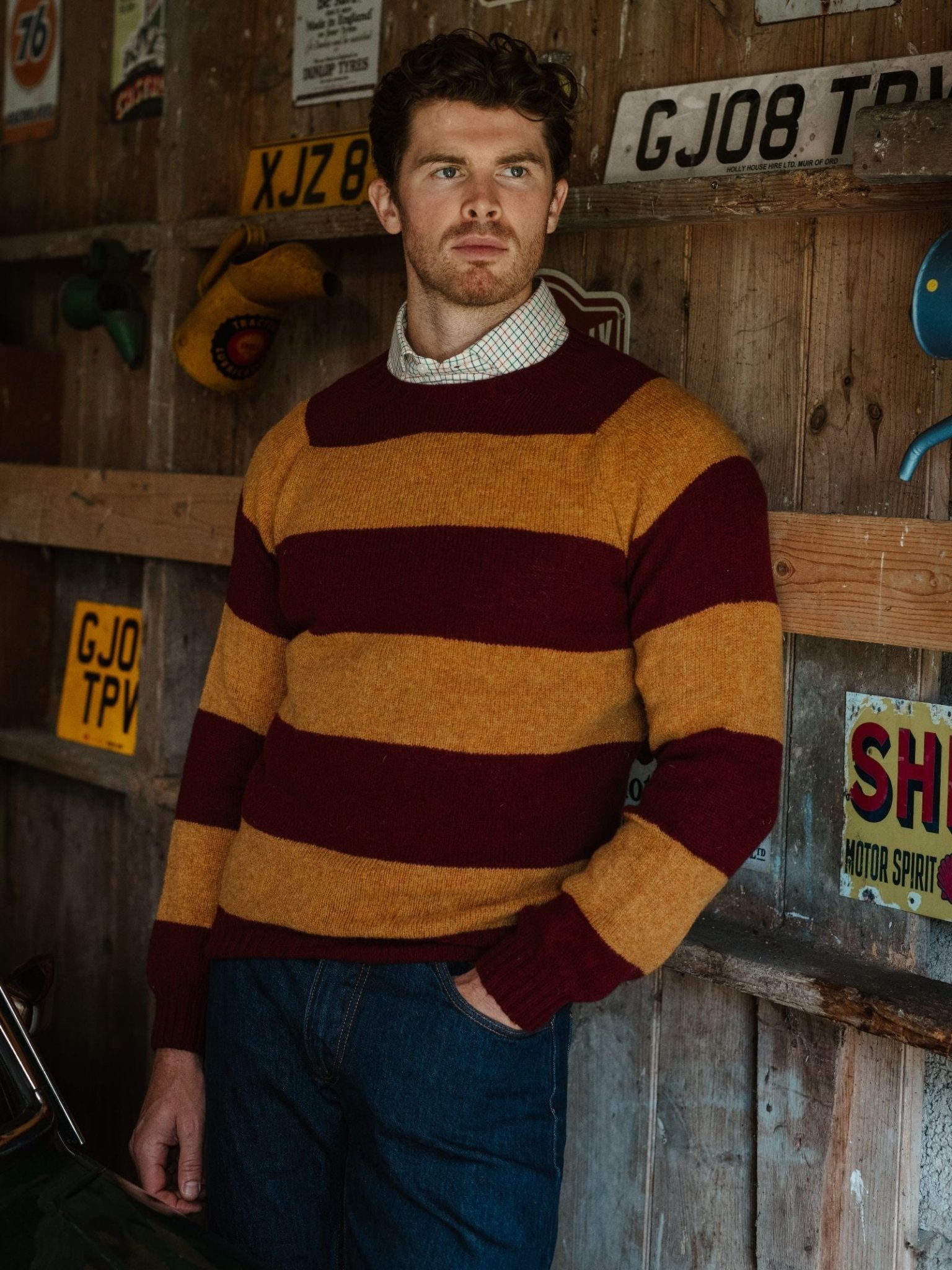 A man with short brown hair and a beard stands in a wood-paneled room decorated with vintage license plates, wearing a Campbell's of Beauly Shetland Stripe Crew Jumper in maroon and mustard over a collared shirt and blue jeans.