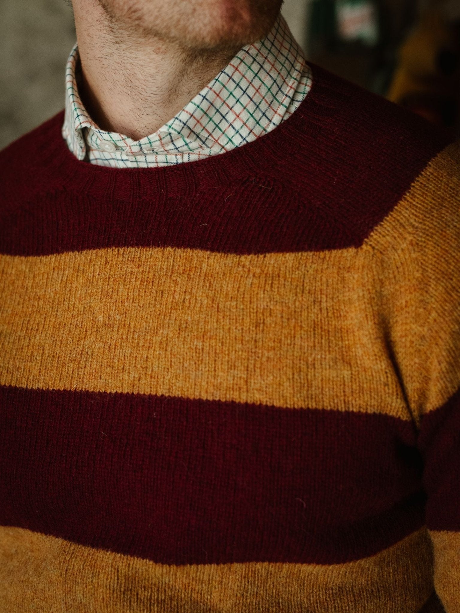 A person wears a maroon and mustard yellow Campbell's of Beauly Shetland Stripe Crew Jumper over a white collared shirt with a green and red check pattern. Only the lower face and upper torso are visible.