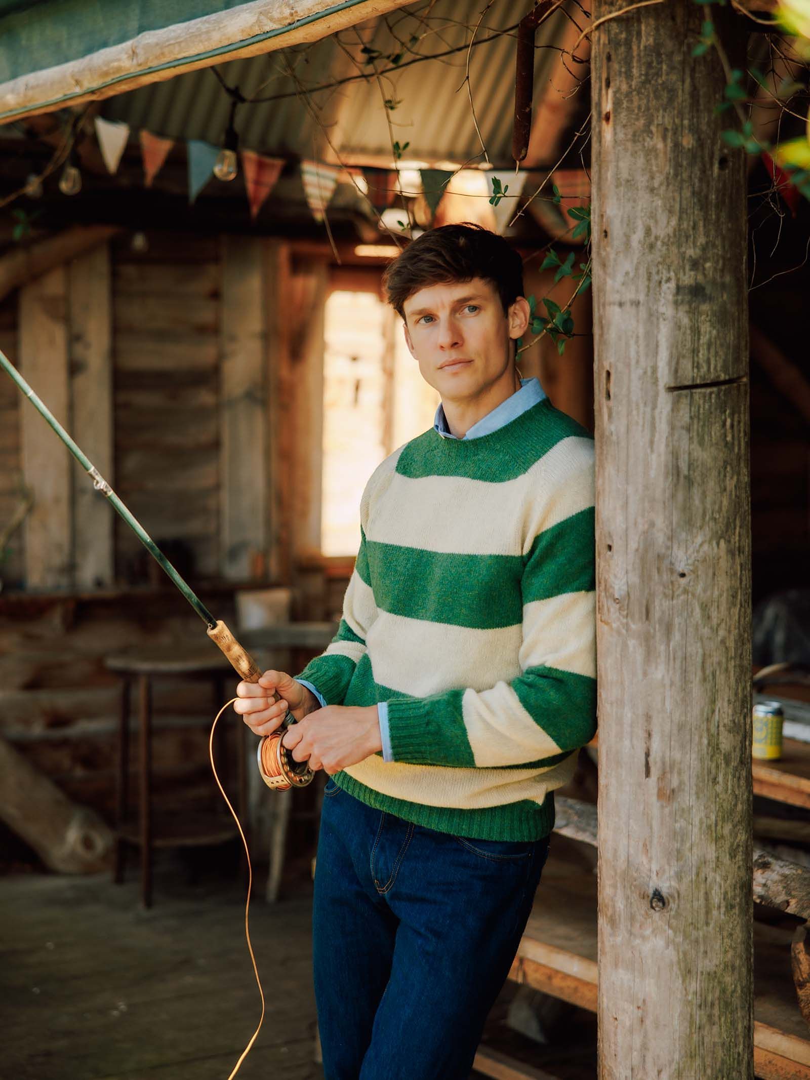 A man wearing the Campbell’s of Beauly Shetland Stripe Crew Jumper, featuring green and white stripes, leans against a wooden post while holding a fishing rod, with a rustic wooden cabin and bunting in the background.