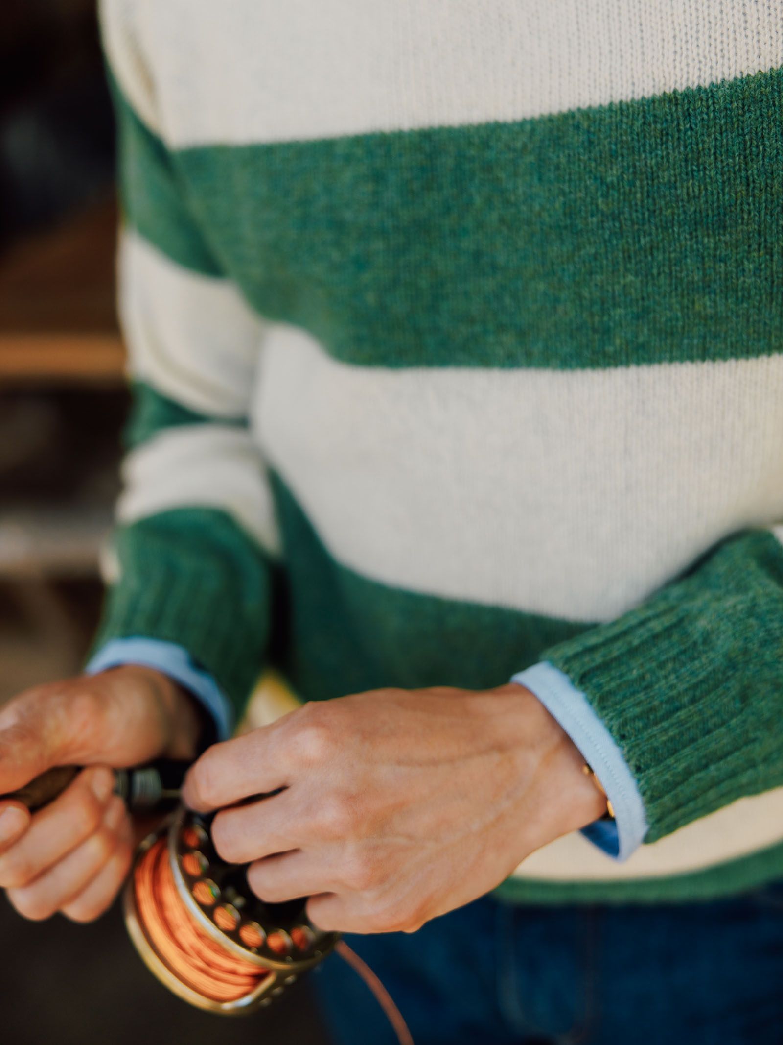 A person wearing the Campbell’s of Beauly Shetland Stripe Crew Jumper holds a fishing reel in their hands, adjusting the line.