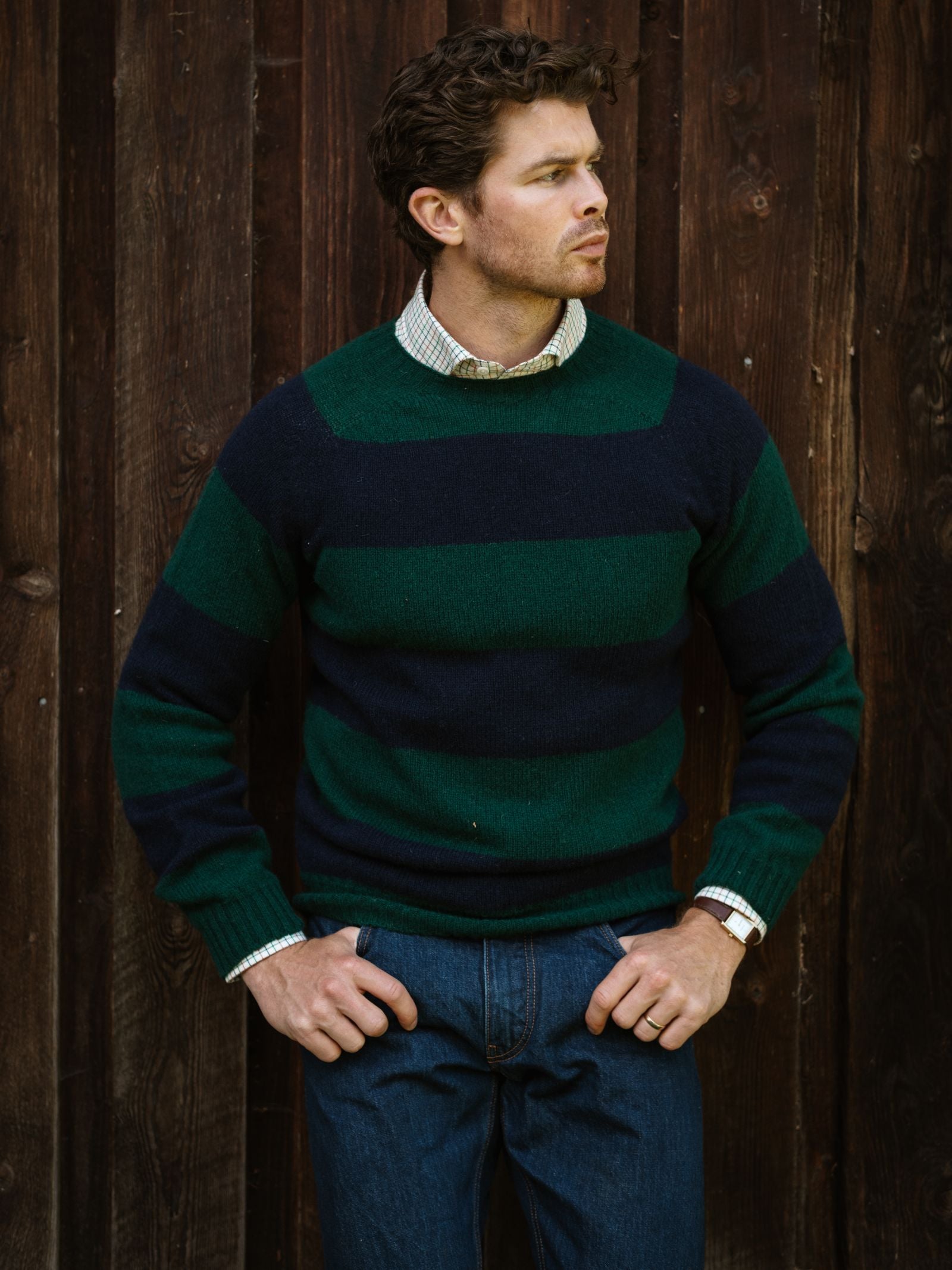 A man wearing the Campbell’s of Beauly Shetland Stripe Crew Jumper and jeans stands before a dark wooden wall, gazing to the side with his hands on his hips.