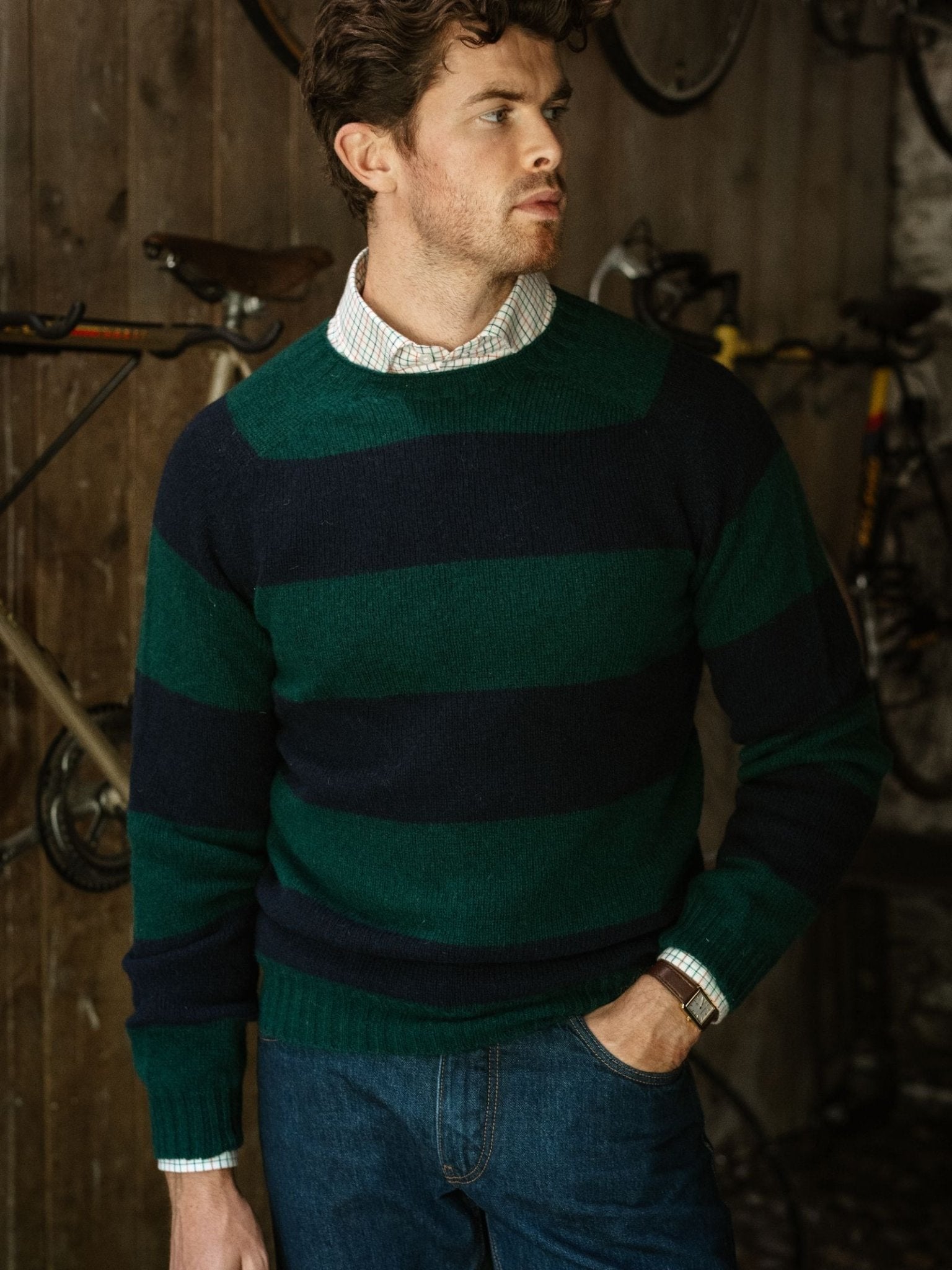 A man wearing the Campbell's of Beauly Shetland Stripe Crew Jumper over a collared shirt stands indoors with his hand in his pocket, looking to the side. Bicycles are mounted on the wooden wall in the background.