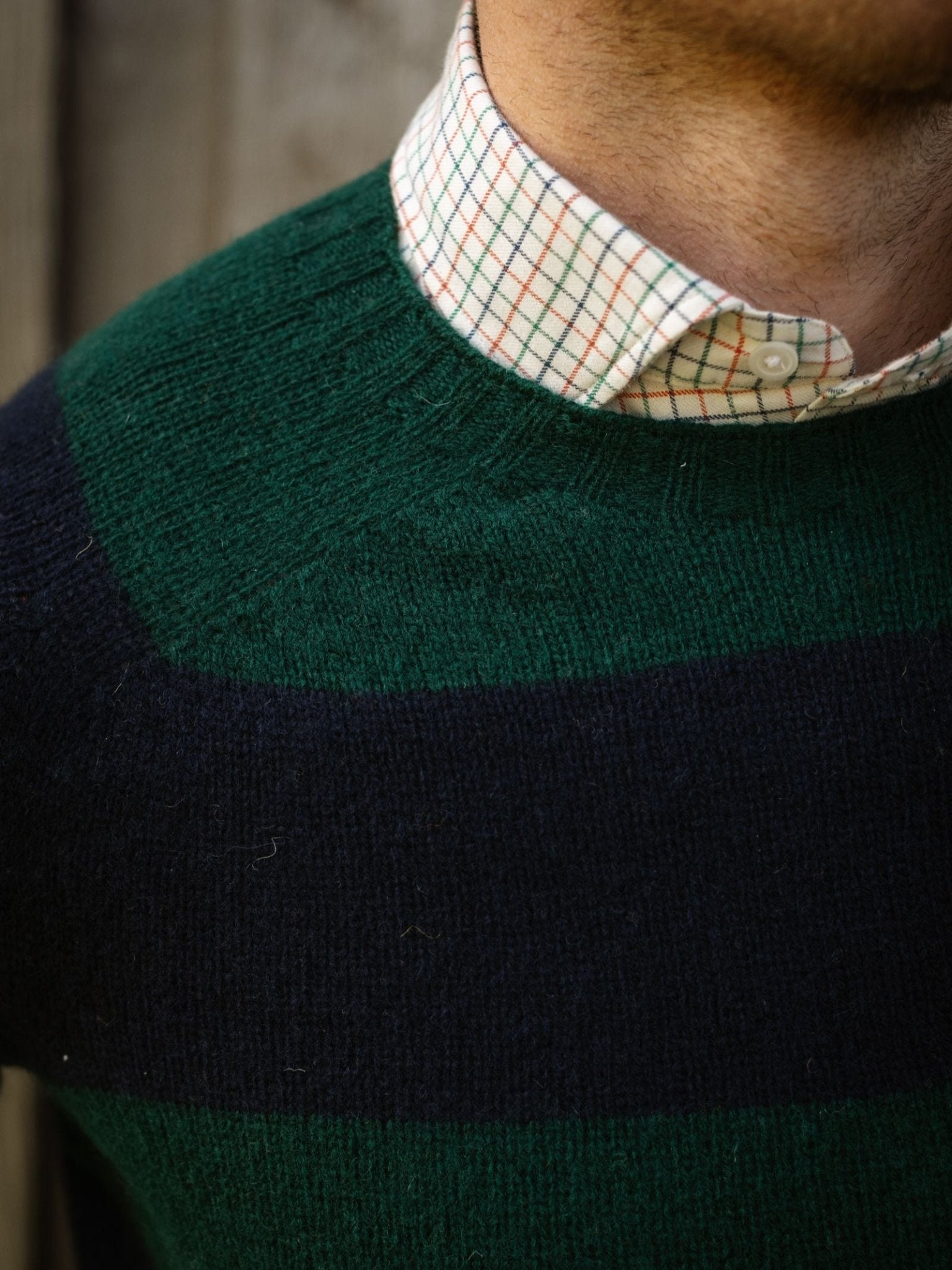 A close-up shows the lower face, neck, and shoulder of a person wearing Campbell's of Beauly Shetland Stripe Crew Jumper—green and navy stripes over a white grid-patterned collared shirt—against a blurred wood background.