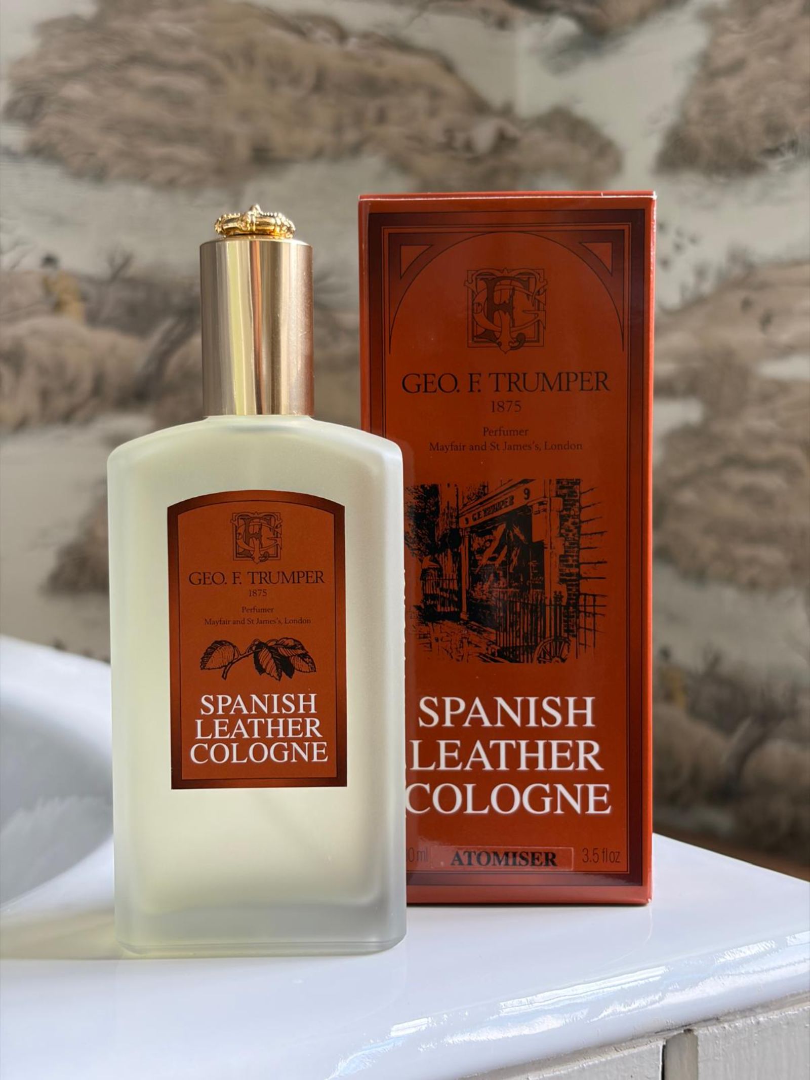A bottle of Geo. F. Trumper Spanish Leather Cologne is displayed before its matching box, both with brown and gold accents, reflecting the musky fragrance against a softly blurred, neutral background.