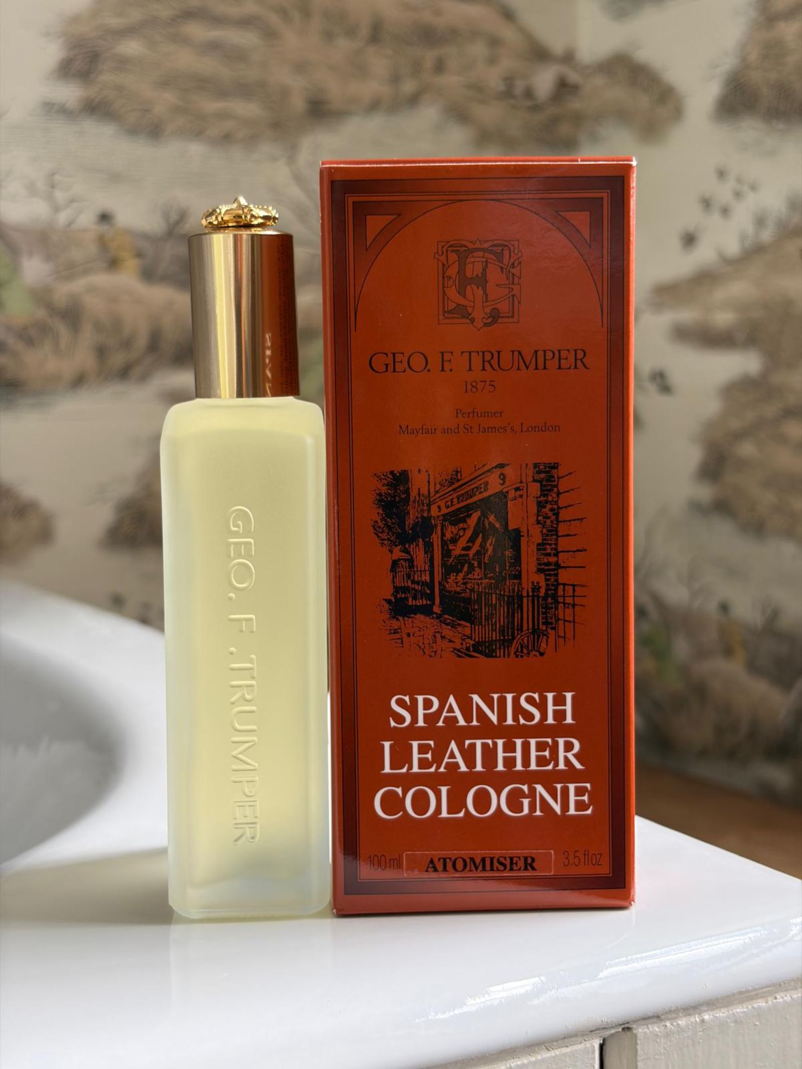 A bottle of Geo. F. Trumper Spanish Leather Cologne sits beside its red and gold box on a white bathroom counter, with patterned wallpaper in the background.
