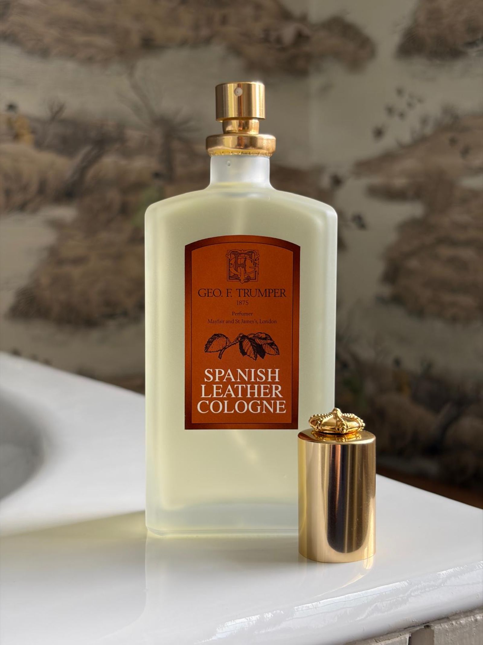 A bottle of Geo. F. Trumper Spanish Leather Cologne rests on a white surface, its gold cap gleaming, while the rich musk scent suggests timeless sophistication against a softly blurred nature-inspired background.