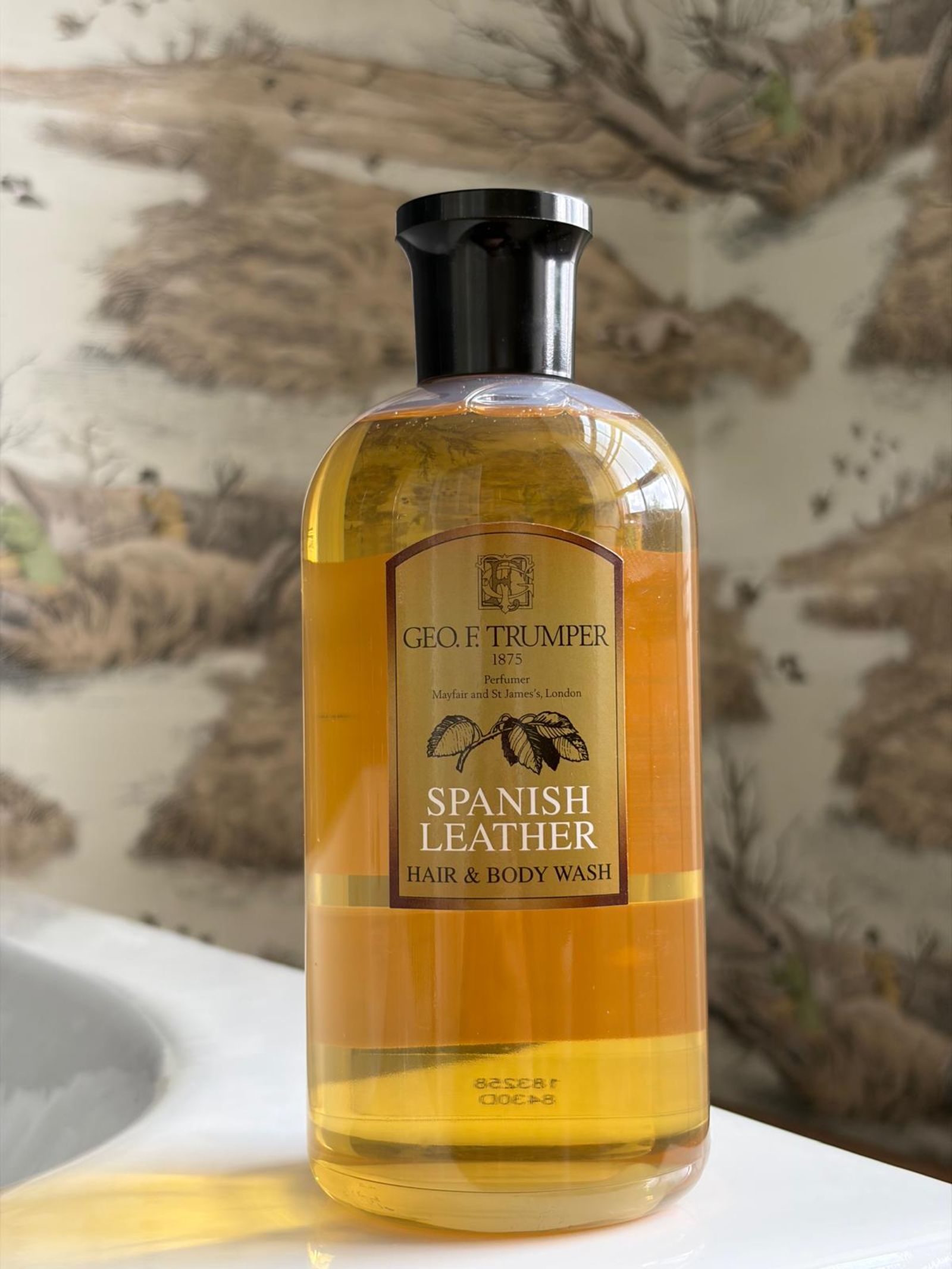 A bottle of Geo. F. Trumper Hair & Body Wash sits on the bathroom counter, its scent filling the air as nature-themed wallpaper with trees and animals decorates the softly blurred background.
