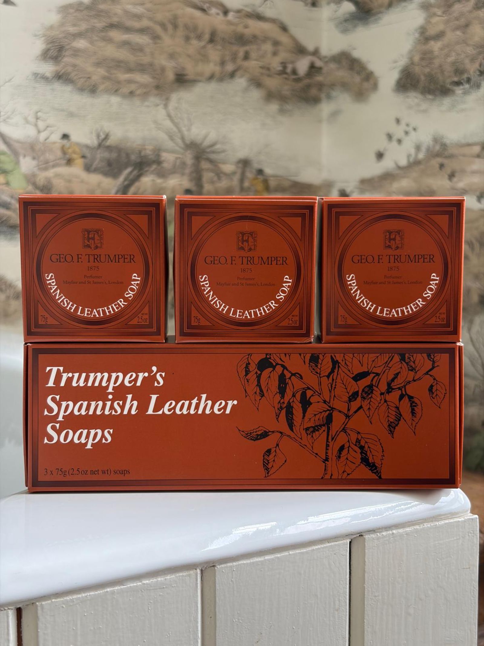 Three orange boxes of Geo. F. Trumper Spanish Leather Hand Soaps, Set of 3, rest on a matching box with decorative text and leaves, displayed on a white surface with a scenic painting in the background.