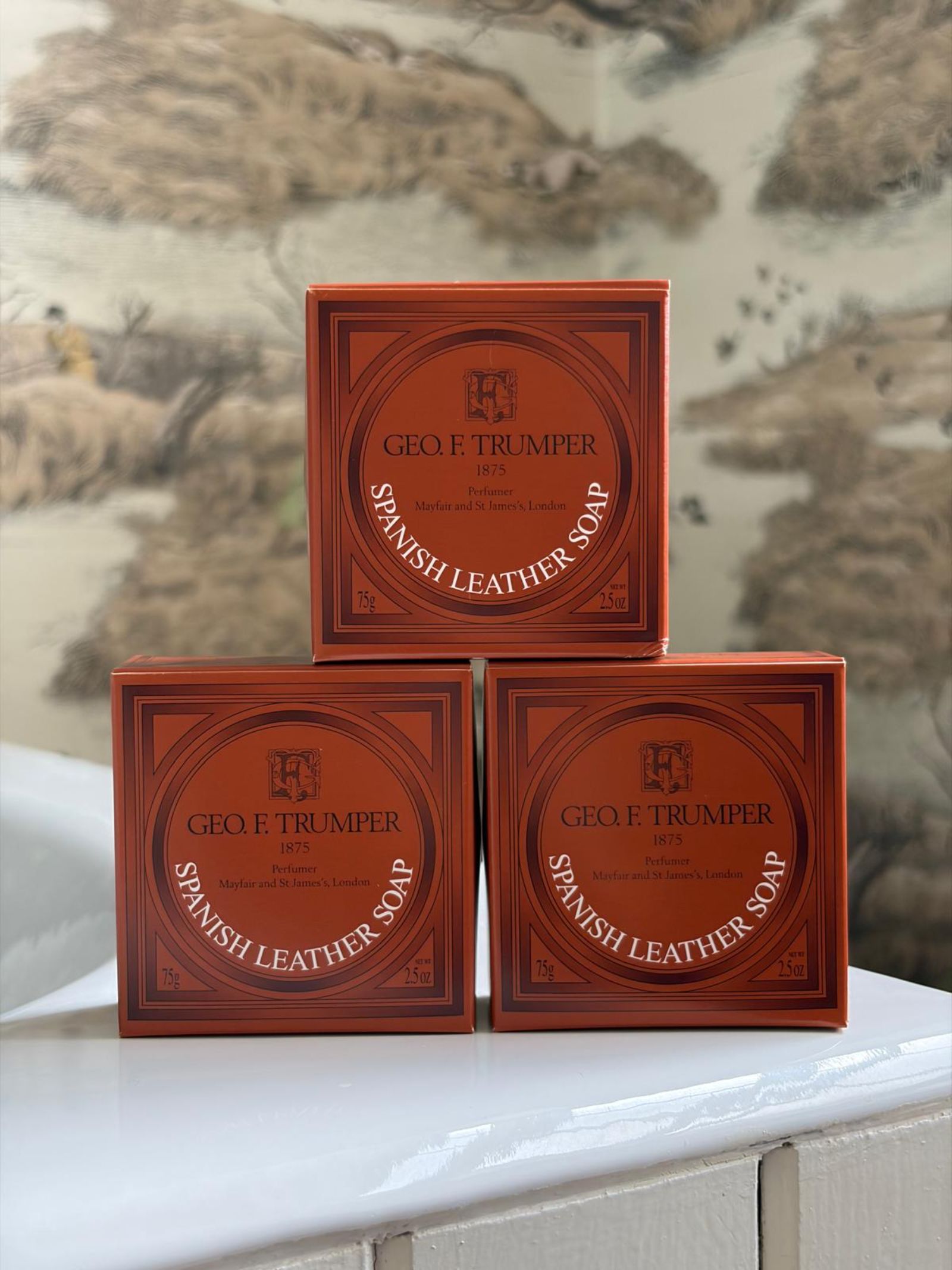 Three orange boxes of Geo. F. Trumper Spanish Leather Hand Soaps, Set of 3, are stacked in a triangle on a white surface against wallpaper with a subtle nature motif.
