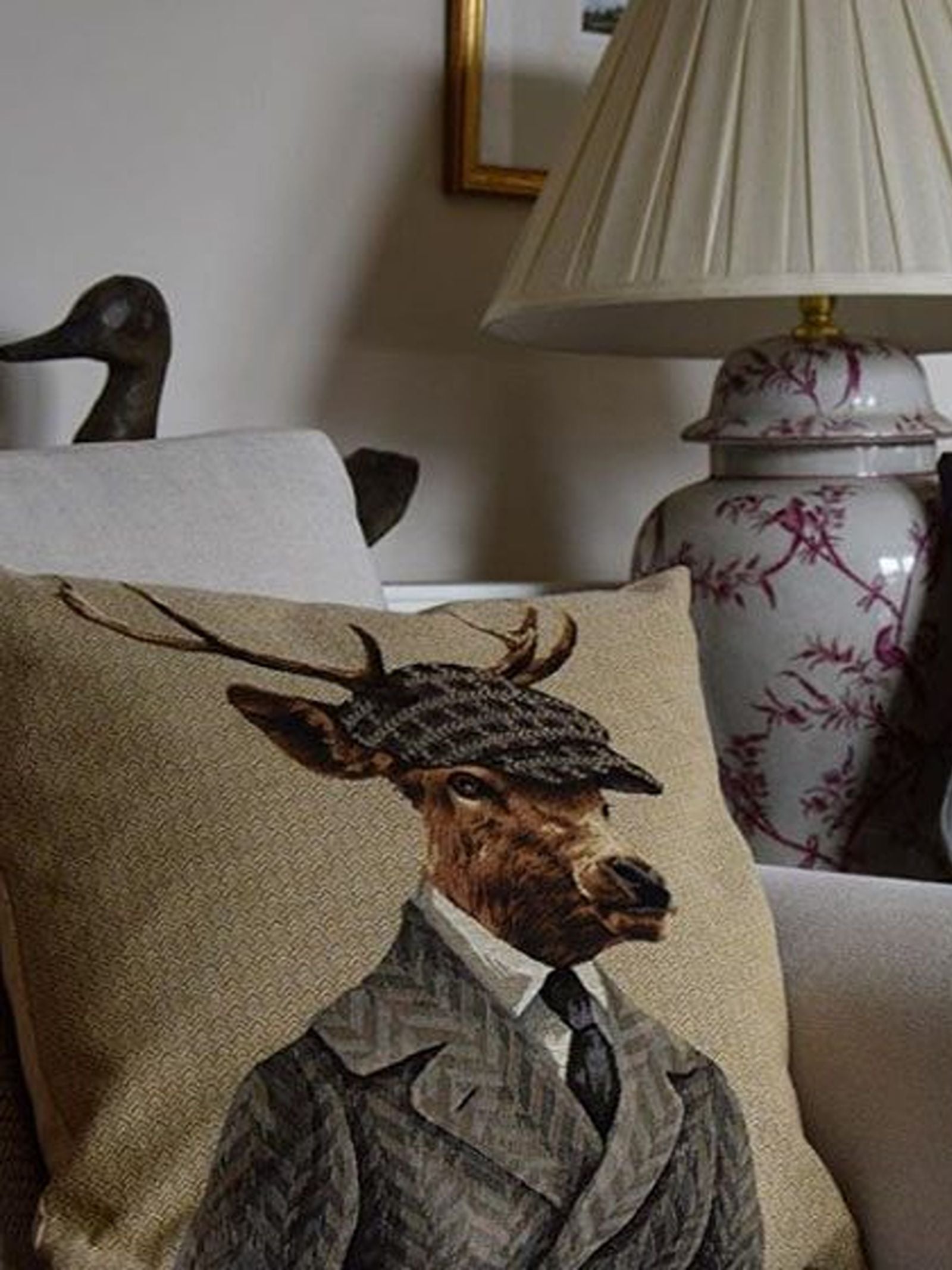 A Campbell’s of Beauly Dapper Stag Cushion Cover rests on a sofa, showcasing a stylish illustration of a deer dressed in a suit and plaid cap. Flanked by a floral table lamp and accompanied by a duck figurine in the background, this unique decorative pillow creates an irresistibly charming scene.