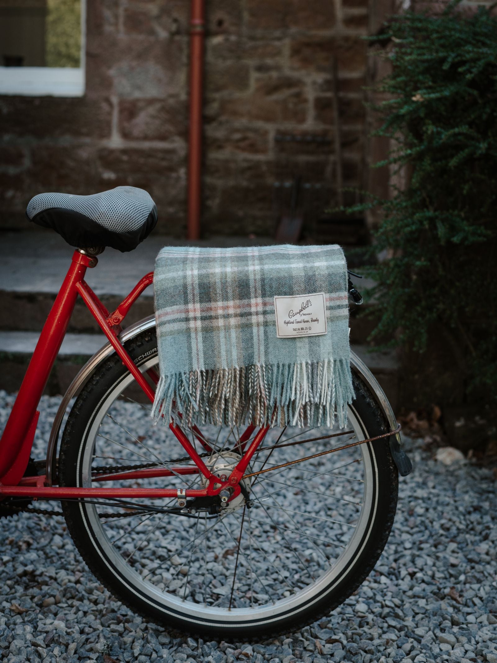 A red bicycle with a gray seat is parked on gravel. A folded blue and gray Strathcarron Lambswool Throw by Campbell's of Beauly, UK made, is draped over the rear wheel. Stone steps and a green bush are in the background.