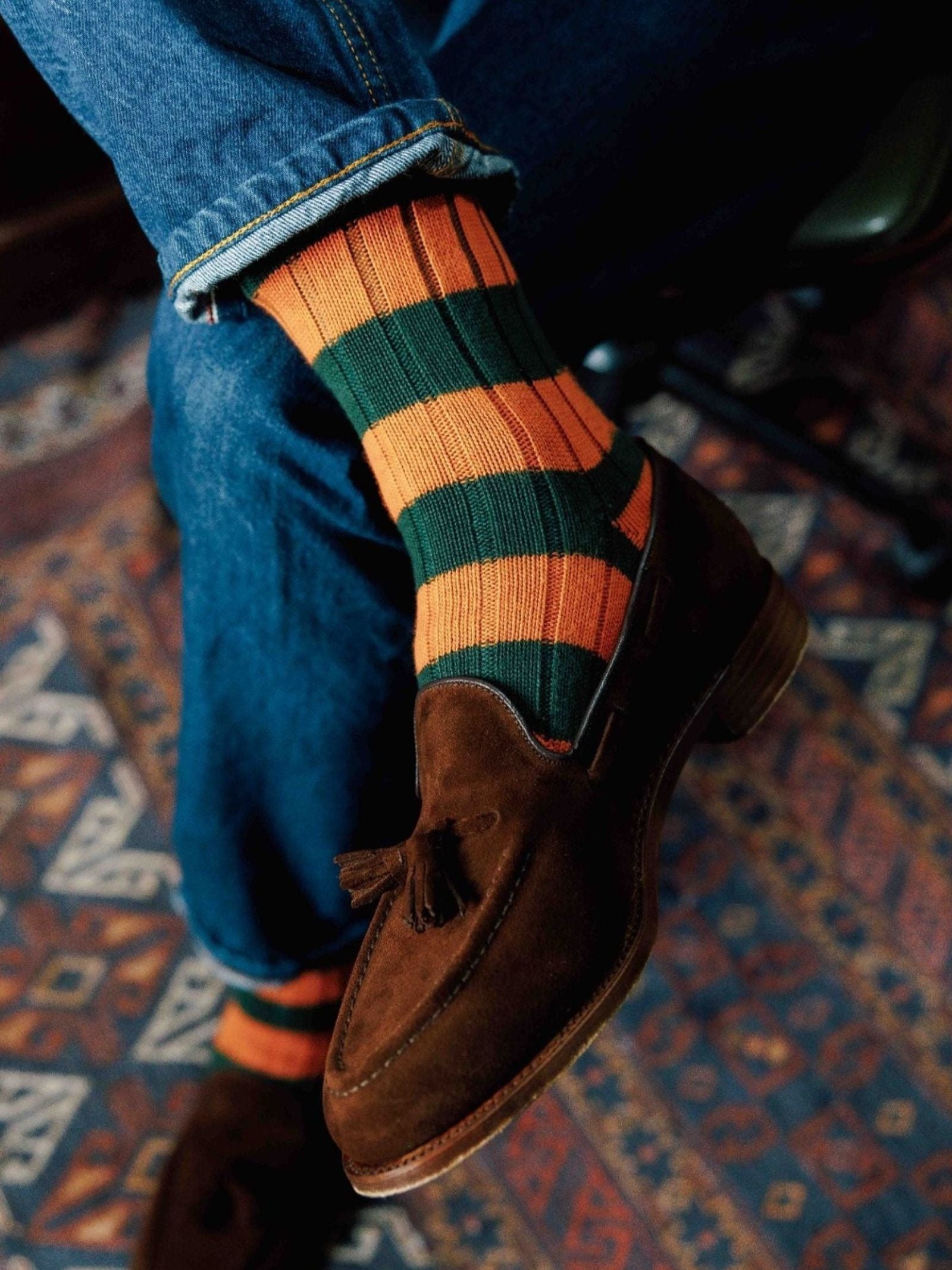 A person sits cross-legged on a patterned rug wearing blue jeans, brown suede tassel loafers, and Campbell's of Beauly Stripe Socks.