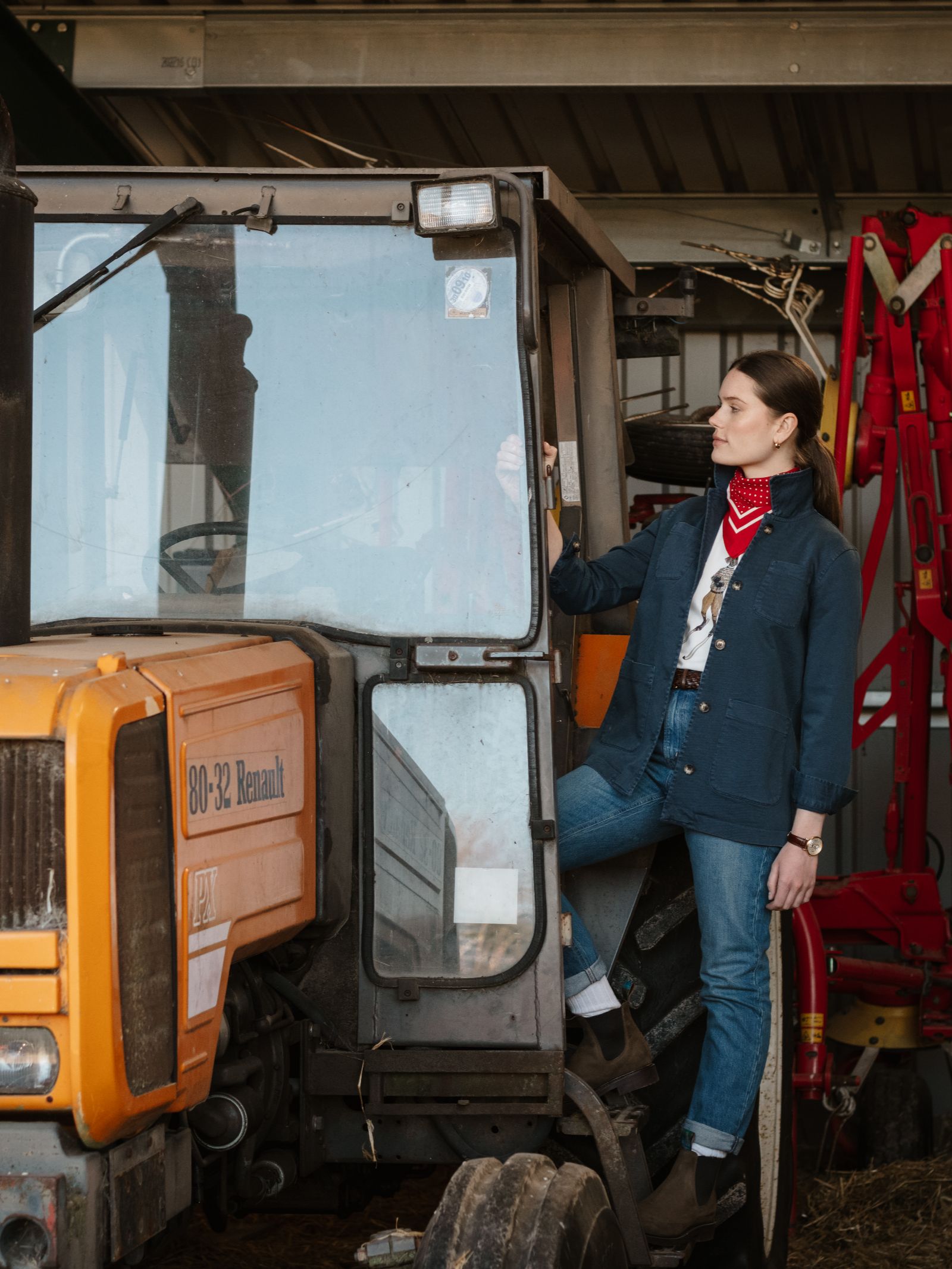 Wearing a red bandana, jeans, and a Campbell's of Beauly Summer Chore Jacket, a person stands with one foot on the step of an orange Renault tractor in a barn, gazing confidently into the distance.