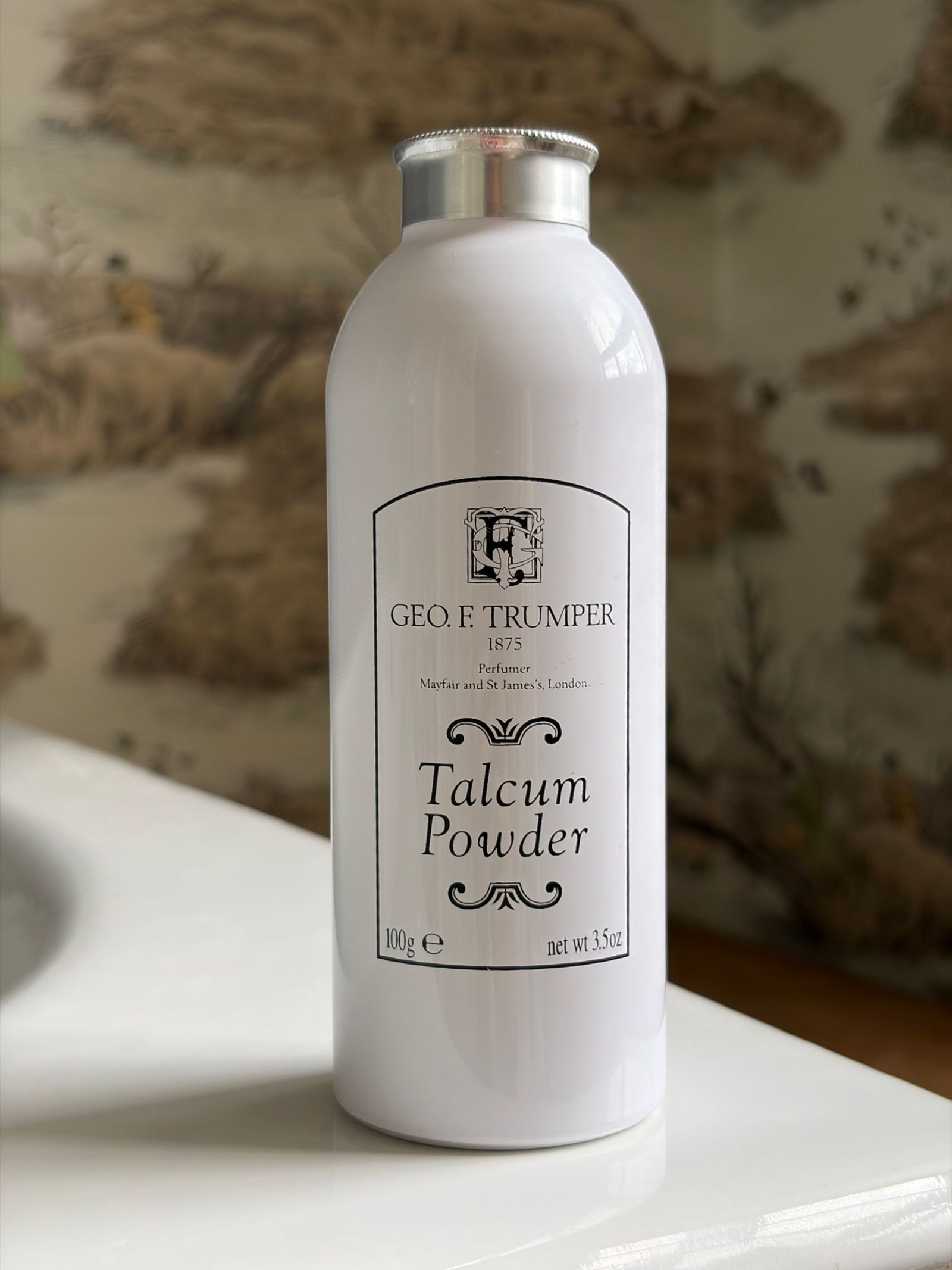 A white bottle of Campbell's of Beauly Talcum Powder with a silver cap sits on a bathroom counter. The label has black text and decorative details, and a blurred patterned wall can be seen in the background.