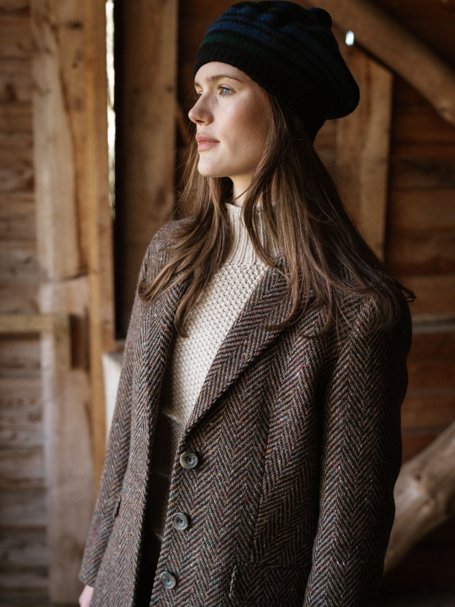 A woman with long brown hair wears the Campbell's of Beauly Lambswool Tartan Beret, a cream sweater, and a brown herringbone coat as she stands indoors, gazing thoughtfully out of frame against a rustic wooden backdrop.
