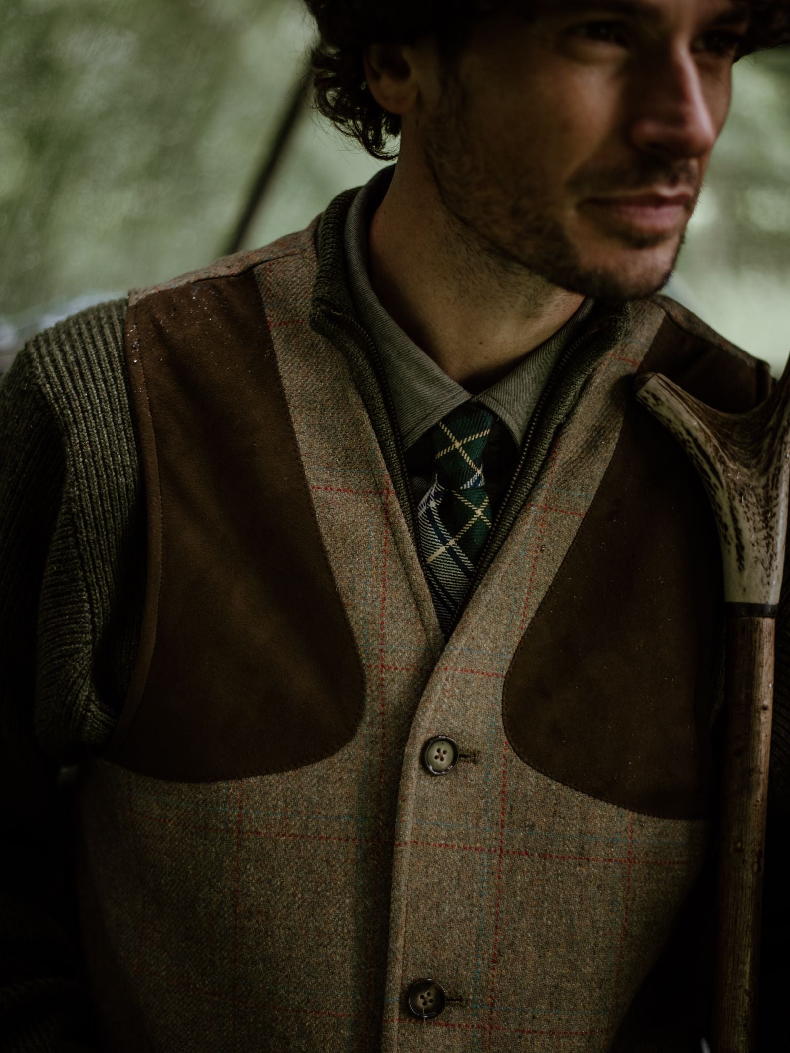 A man wears a Campbell's of Beauly Tartan Tie with a collared shirt and brown tweed vest outdoors, softly lit from the shoulders up as he looks to the side and holds an antler.
