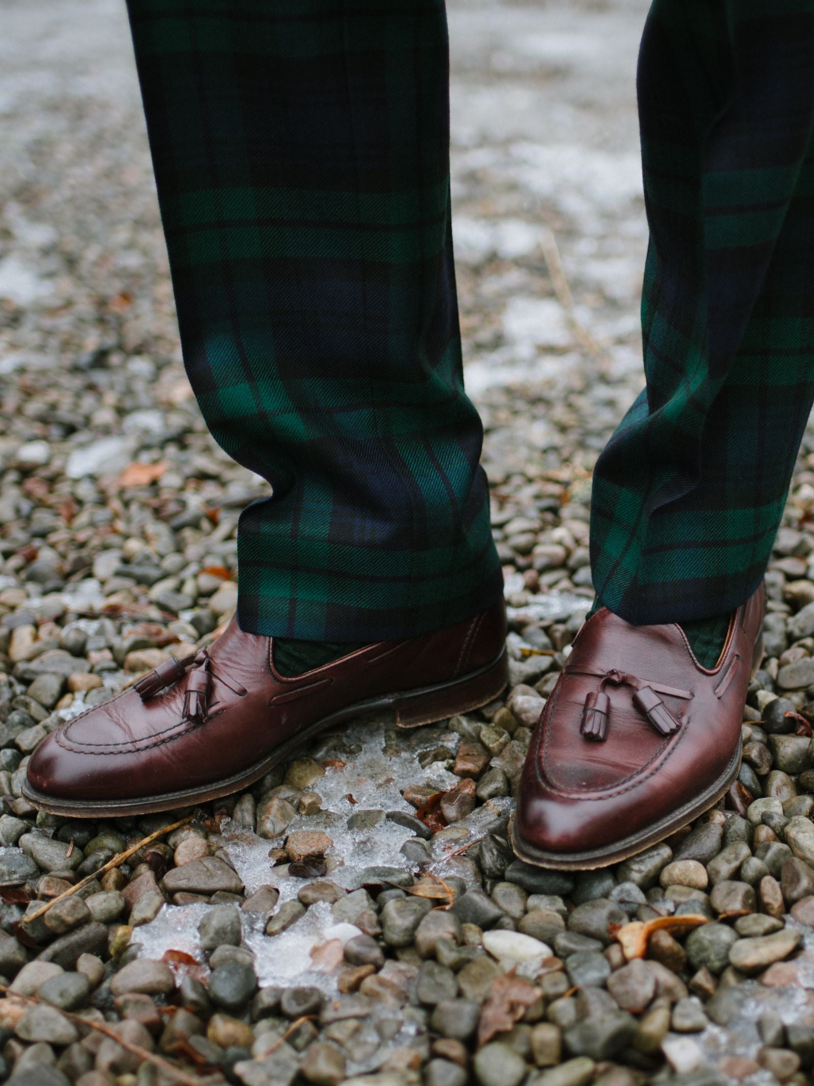A person wearing Campbell's of Beauly Auld Stock Tartan Trousers—100% wool, made in the UK—and brown leather tassel loafers stands on gravel and patches of ice.