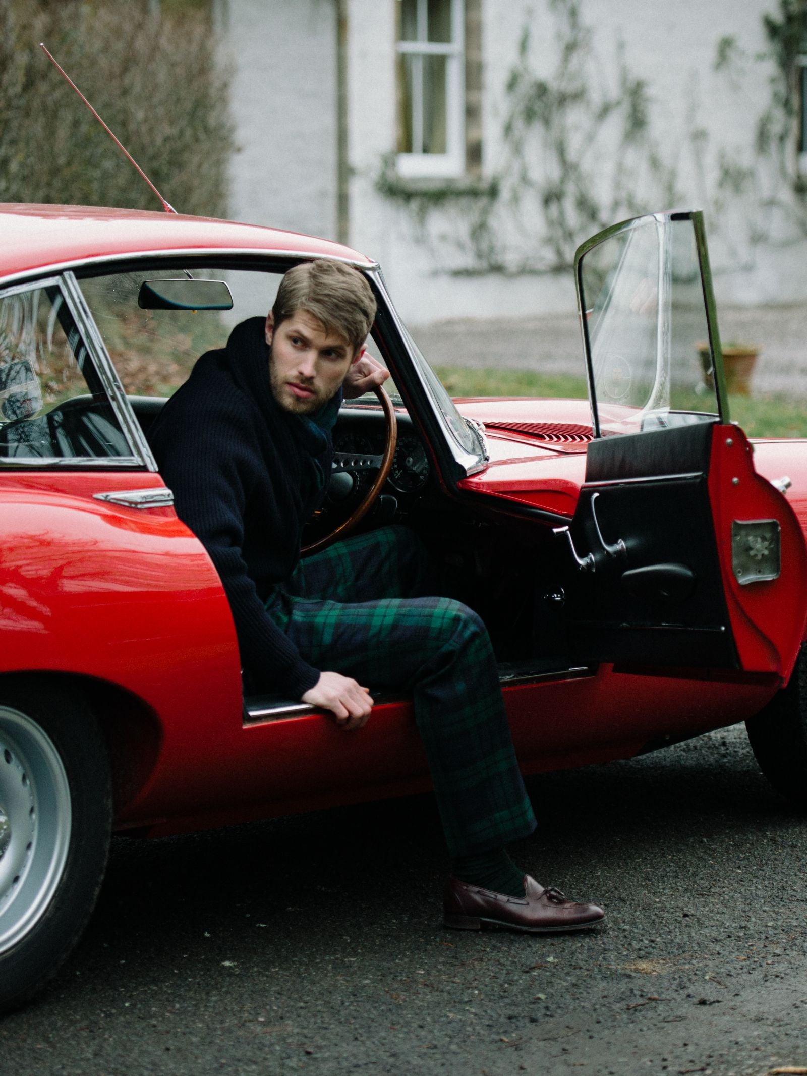 A man in a dark sweater and Campbell's of Beauly Auld Stock Tartan Trousers in Blackwatch sits in the driver’s seat of a classic red car with the door open, looking out and holding the steering wheel. Blurred building and plants appear in the background.