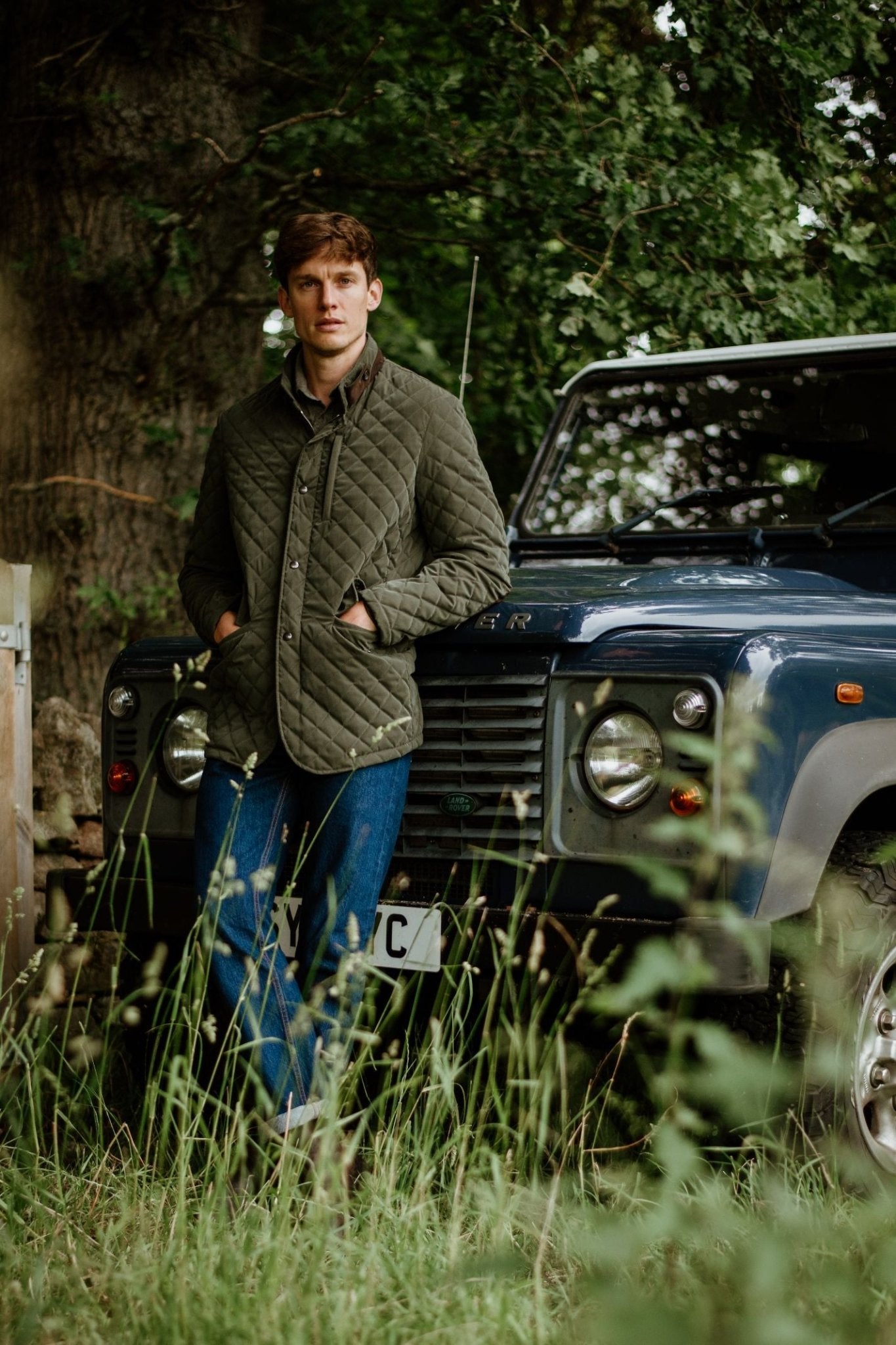 Wearing the Campbell's of Beauly Auld Stock Tomich Jacket and blue jeans, a person stands outdoors by a dark vintage off-road vehicle amid tall grass and greenery, highlighting timeless country style.