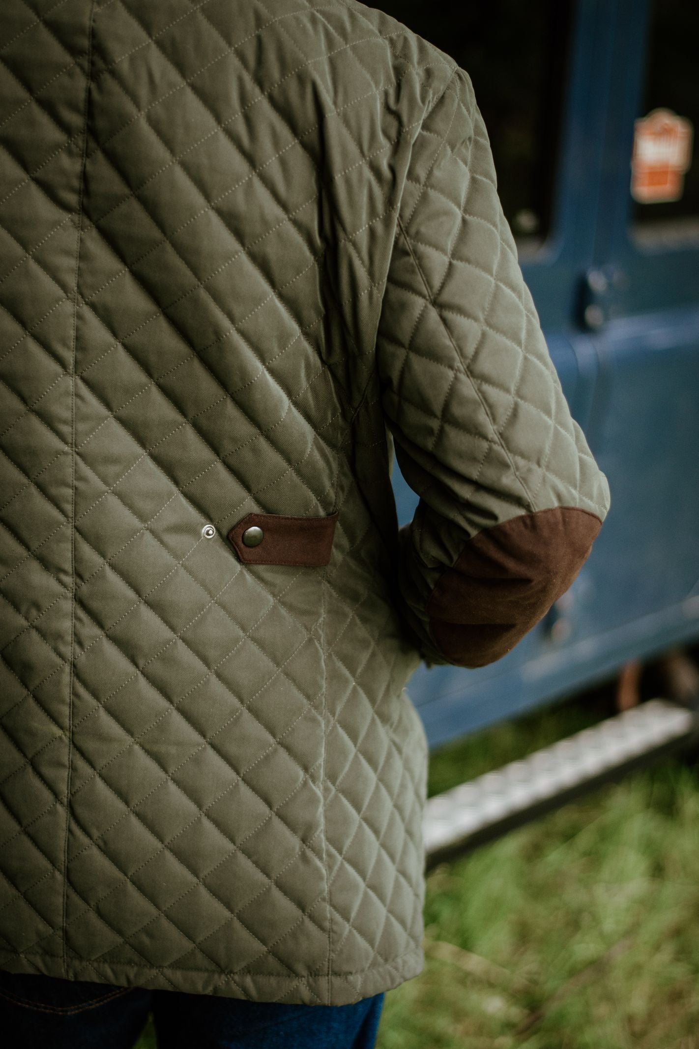 Wearing the Auld Stock Tomich Jacket by Campbell's of Beauly, a person stands outdoors by a blue vehicle. Only their back and arm with brown elbow patch are visible, against grass at the bottom of the image.
