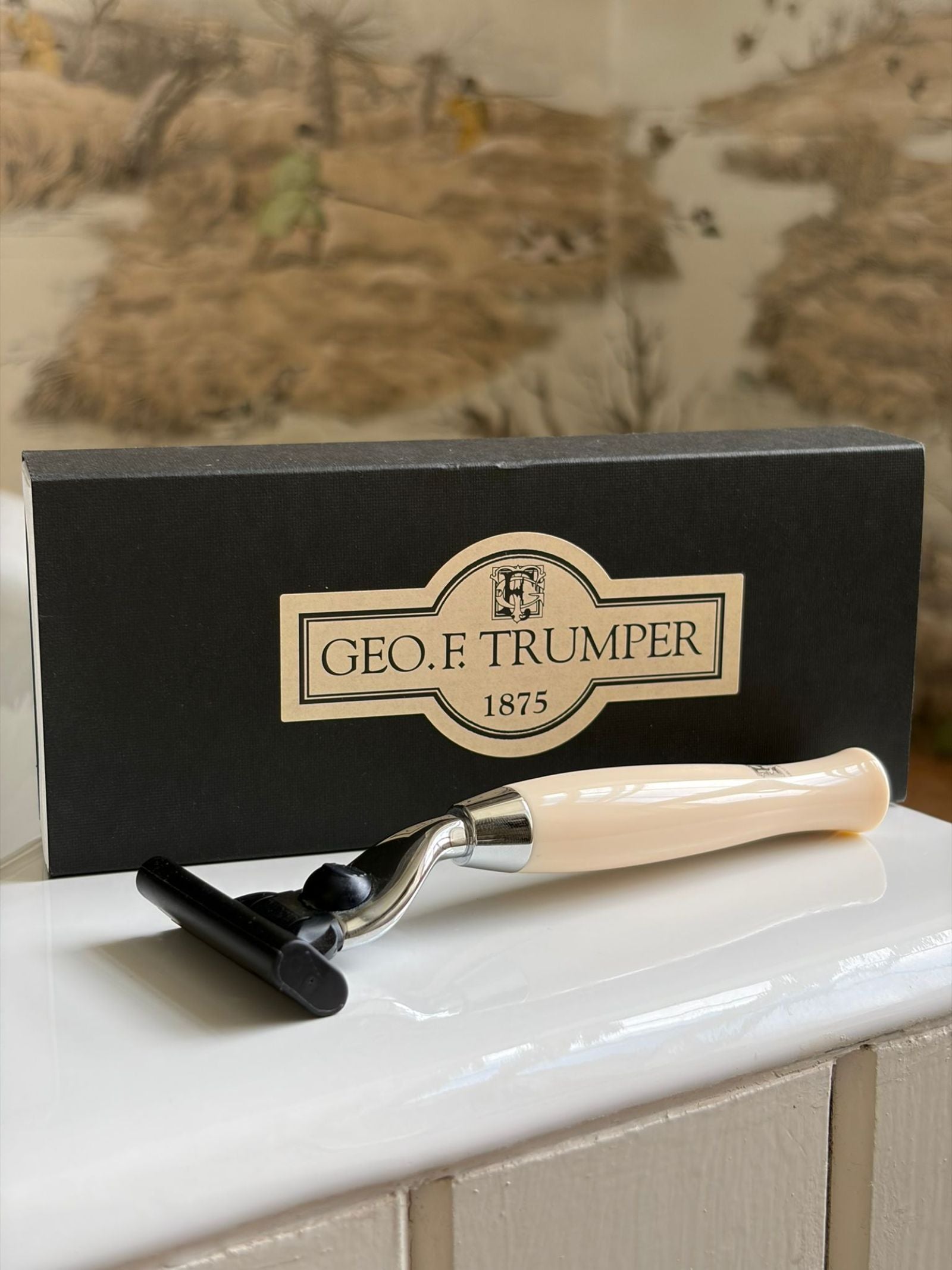 A Geo. F. Trumper Simulated Ivory Mach 3 Razor Handle rests in front of a black Geo. F. Trumper 1875 box on a white surface, with scenic wallpaper in the background. Compatible with Mach 3 blades.