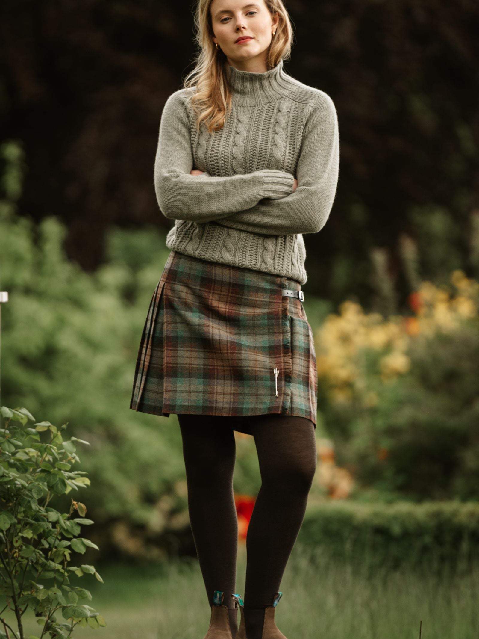 A woman poses outdoors among greenery in a Campbell's of Beauly Tweed Kilt, paired with a grey cable knit sweater, black tights, and brown boots.