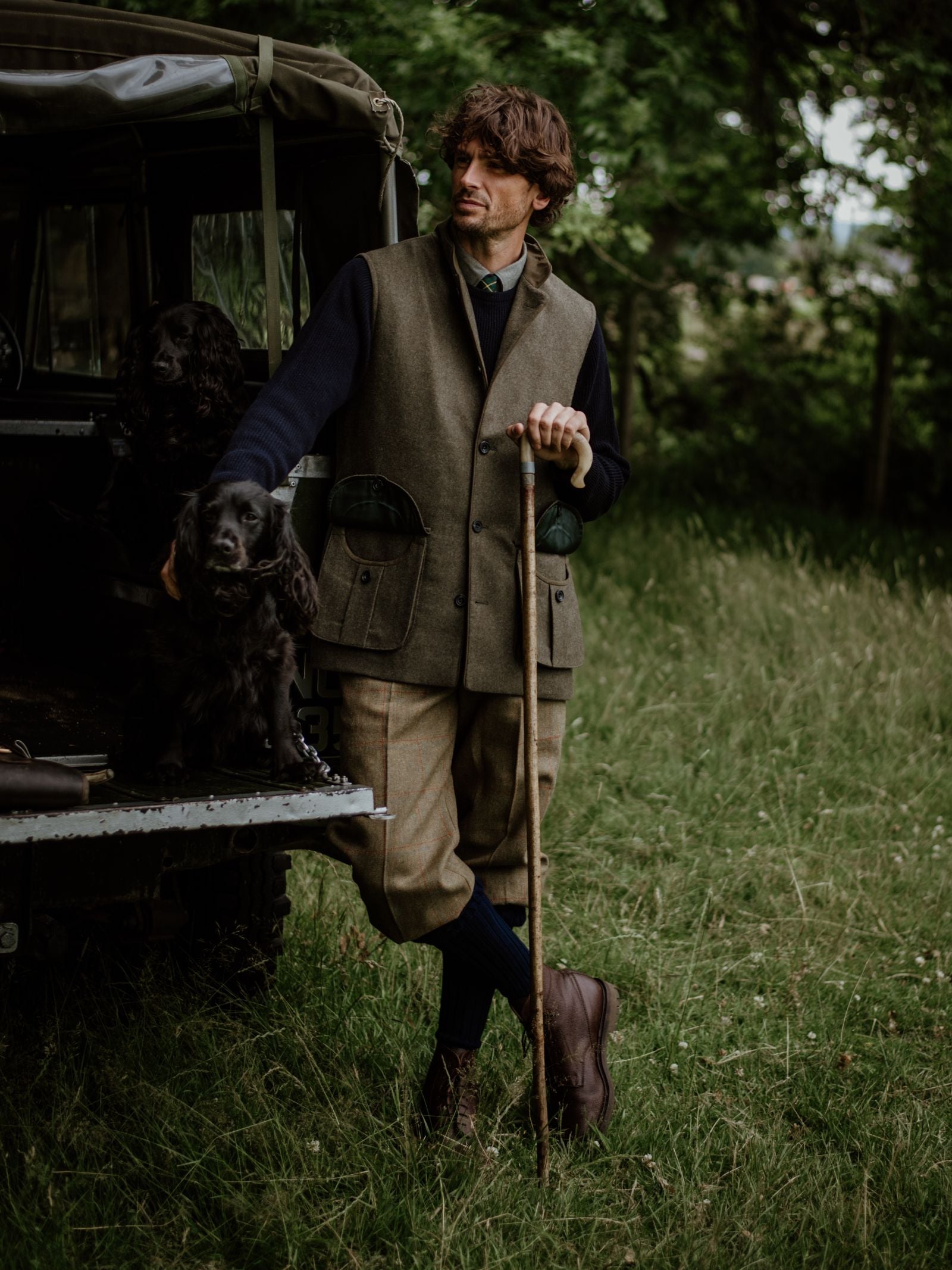 A man in Campbell's of Beauly Plus Fours stands in a grassy field beside an open vehicle, petting two black dogs on the tailgate, his classic country attire blending with the surrounding landscape and trees.