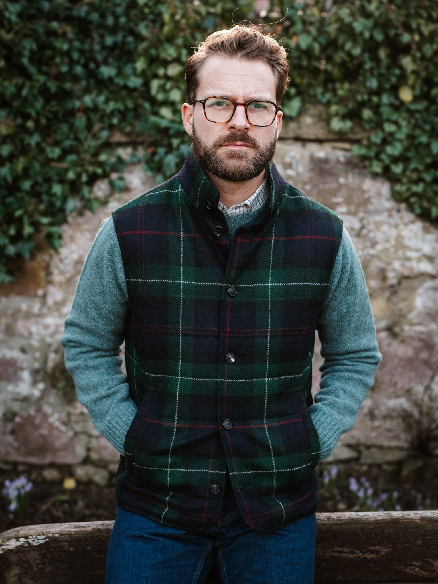 A man wears the Campbell's of Beauly Auld Stock Quilted Tweed Buttoned Gilet and glasses over a gray sweater, standing outdoors by an ivy-clad stone wall, hands in pockets—a timeless layering piece for cool weather.