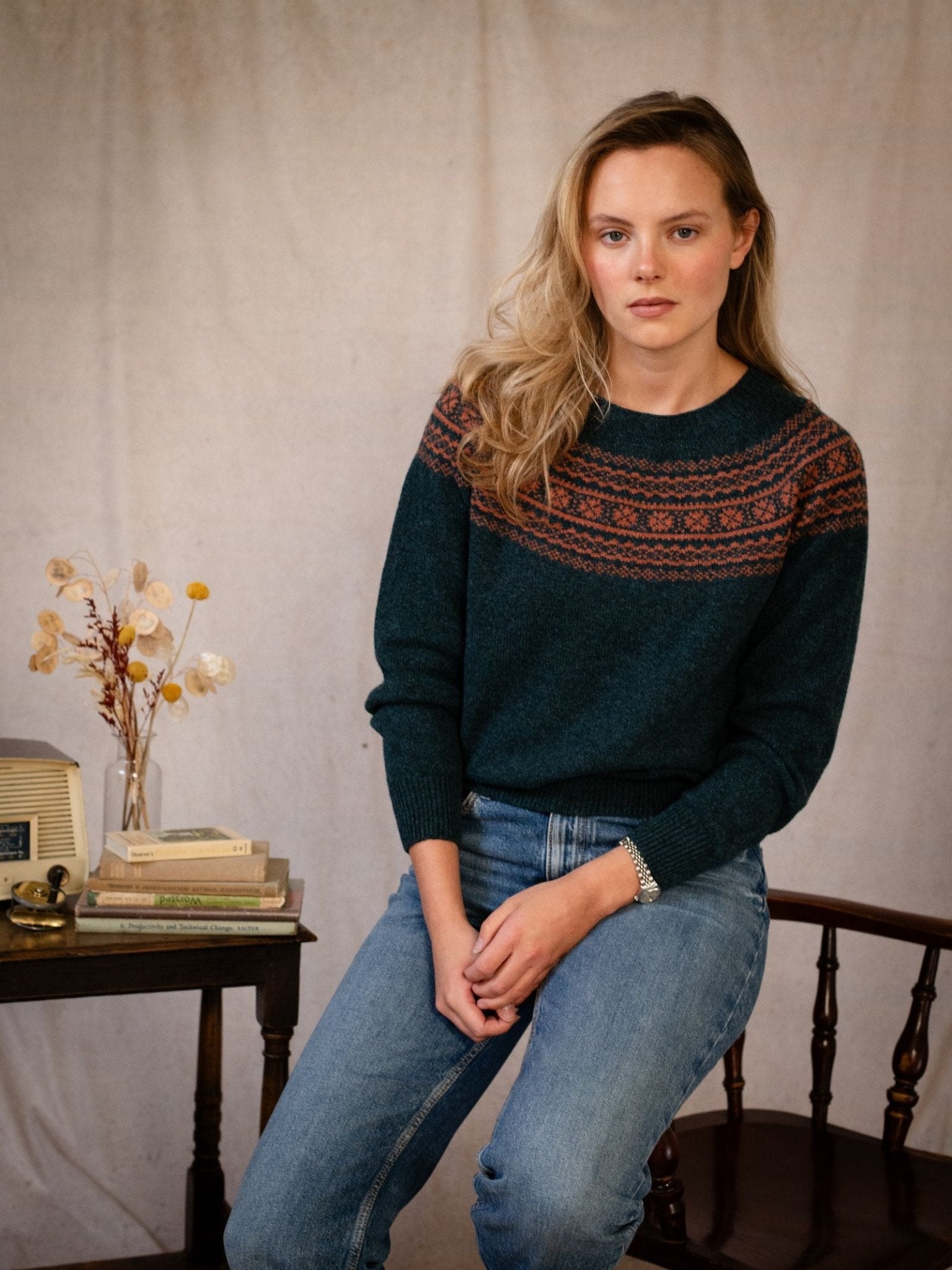 A woman with long blonde hair sits on a chair in jeans, wearing the Campbell's of Beauly Two-Colour Fairisle Yoke Jumper. Next to her is a small table with books, glasses, and a vase of dried flowers against a neutral background.