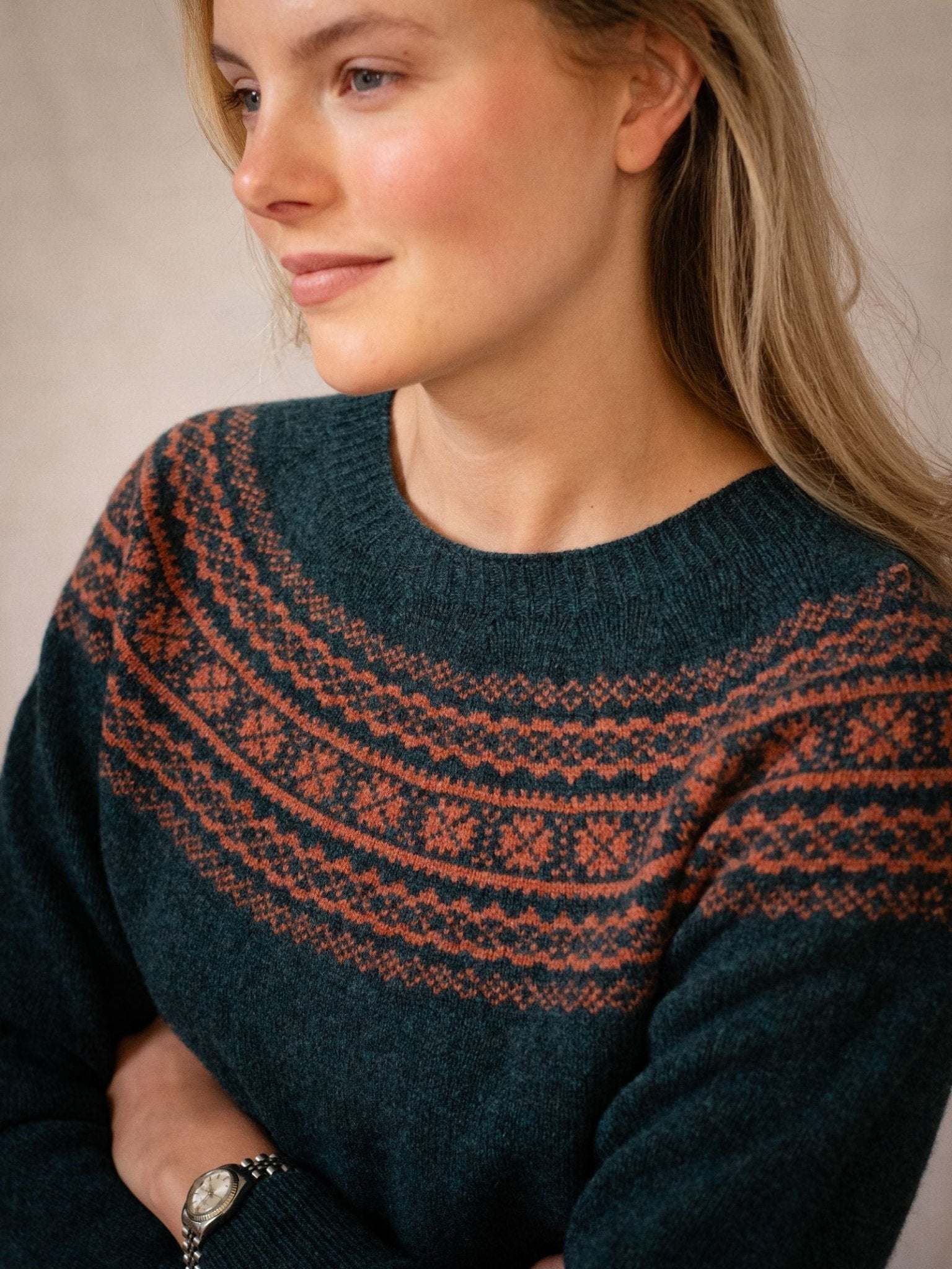 A woman with long blonde hair and fair skin wears the Campbell's of Beauly Two-Colour Fairisle Yoke Jumper in dark green, featuring a red and orange yoke pattern. She stands arms crossed, looking to the side with a slight smile.
