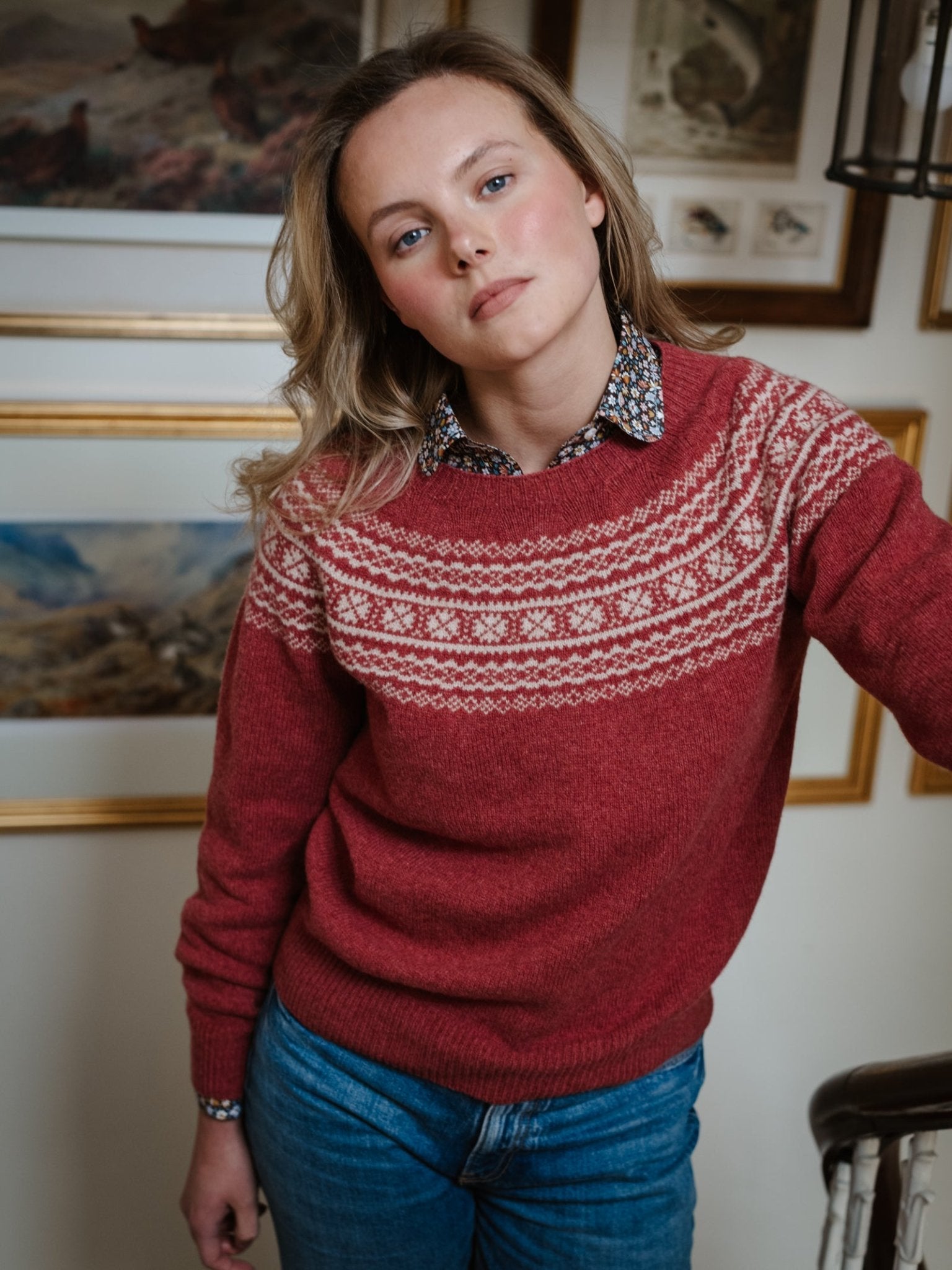 A woman with wavy blonde hair wears the Campbell's of Beauly Two-Colour Fairisle Yoke Jumper and blue jeans, leaning on an indoor staircase railing with framed art pieces visible behind her.