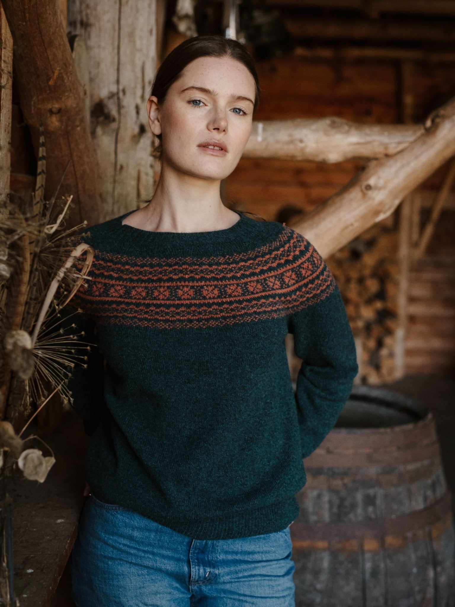 A woman with light skin and brown hair stands indoors, wearing a green Two-Colour Fairisle Yoke Jumper by Campbell's of Beauly and blue jeans. She is in front of rustic wooden beams, a barrel, and dried plants.