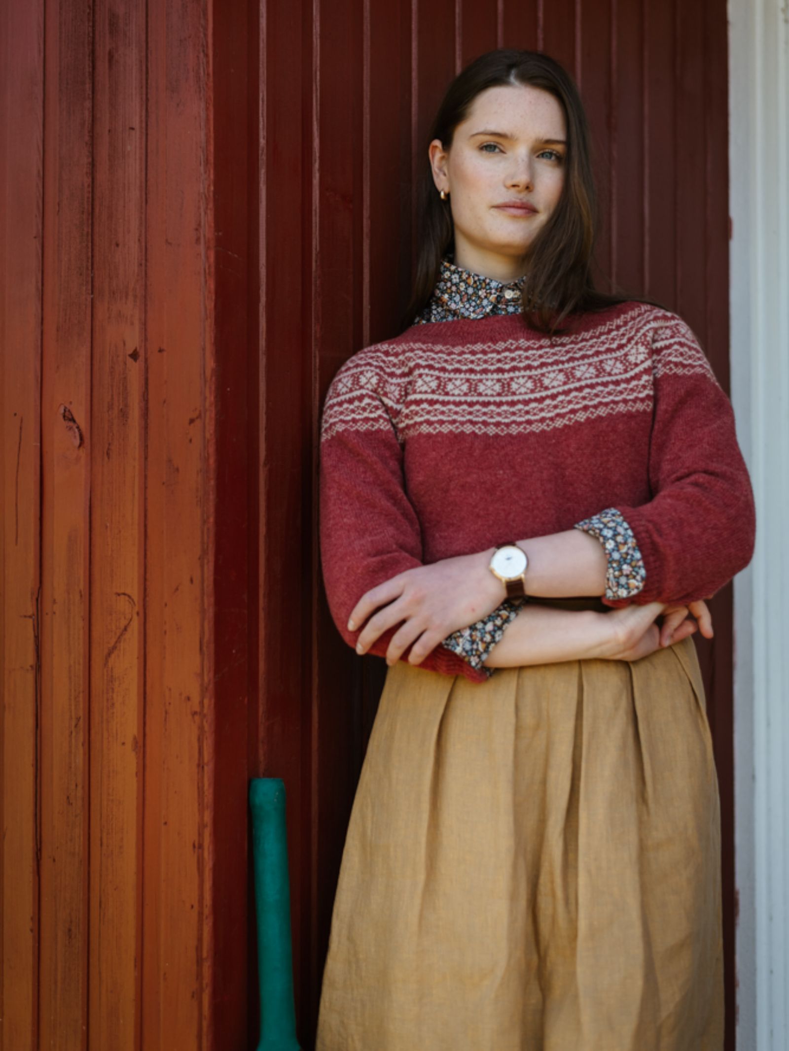 A woman with long brown hair stands before a red wooden wall, arms crossed, wearing Campbell's of Beauly's Two-Colour Fairisle Yoke Jumper over a floral blouse and a light brown skirt, with a watch on her wrist.