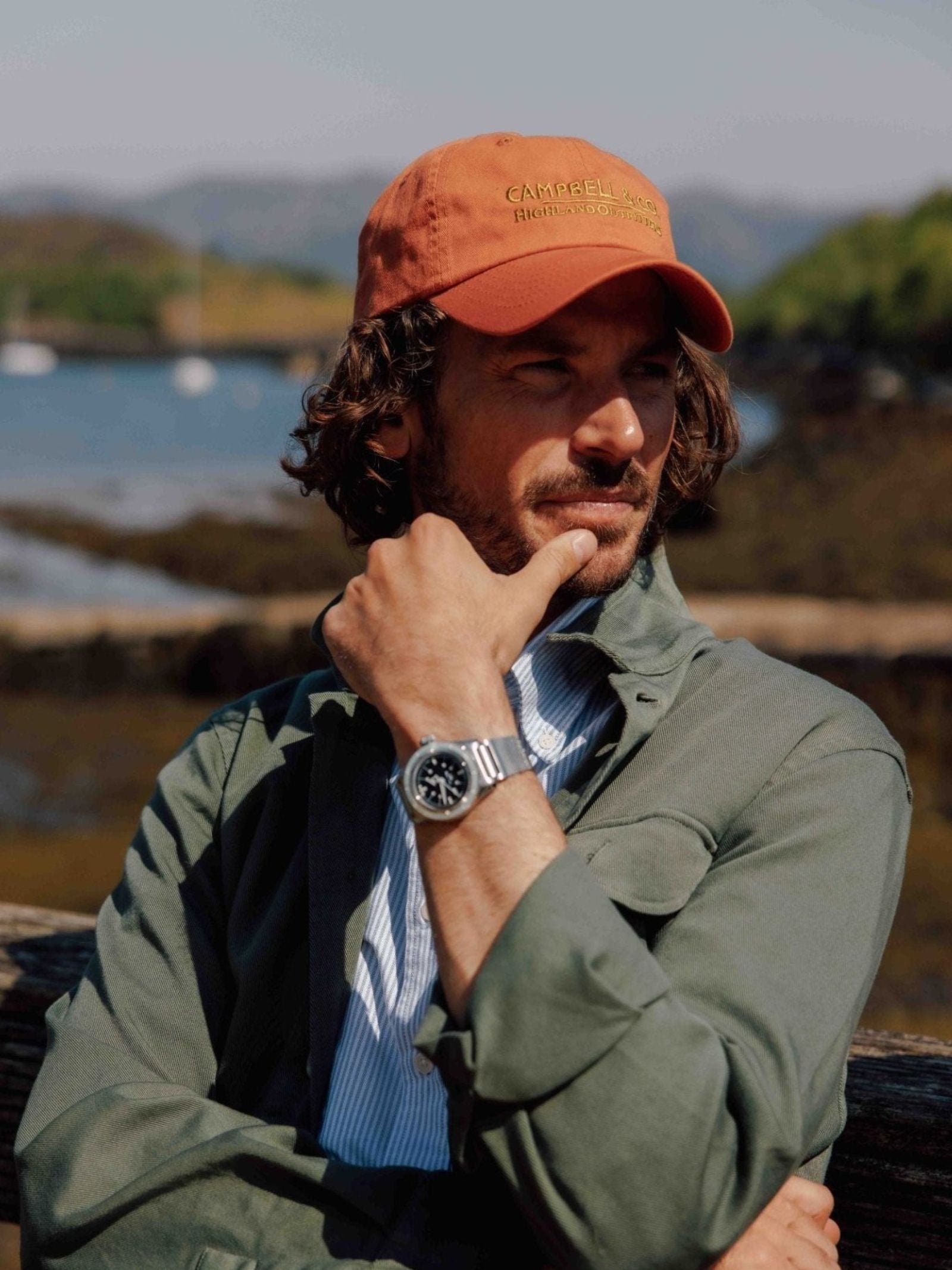 A man with wavy hair wears the Unisex Campbell's Baseball Cap by Campbell's of Beauly, paired with a green jacket and watch, as he rests his chin on his hand and gazes thoughtfully across water and mountains—effortless everyday style.