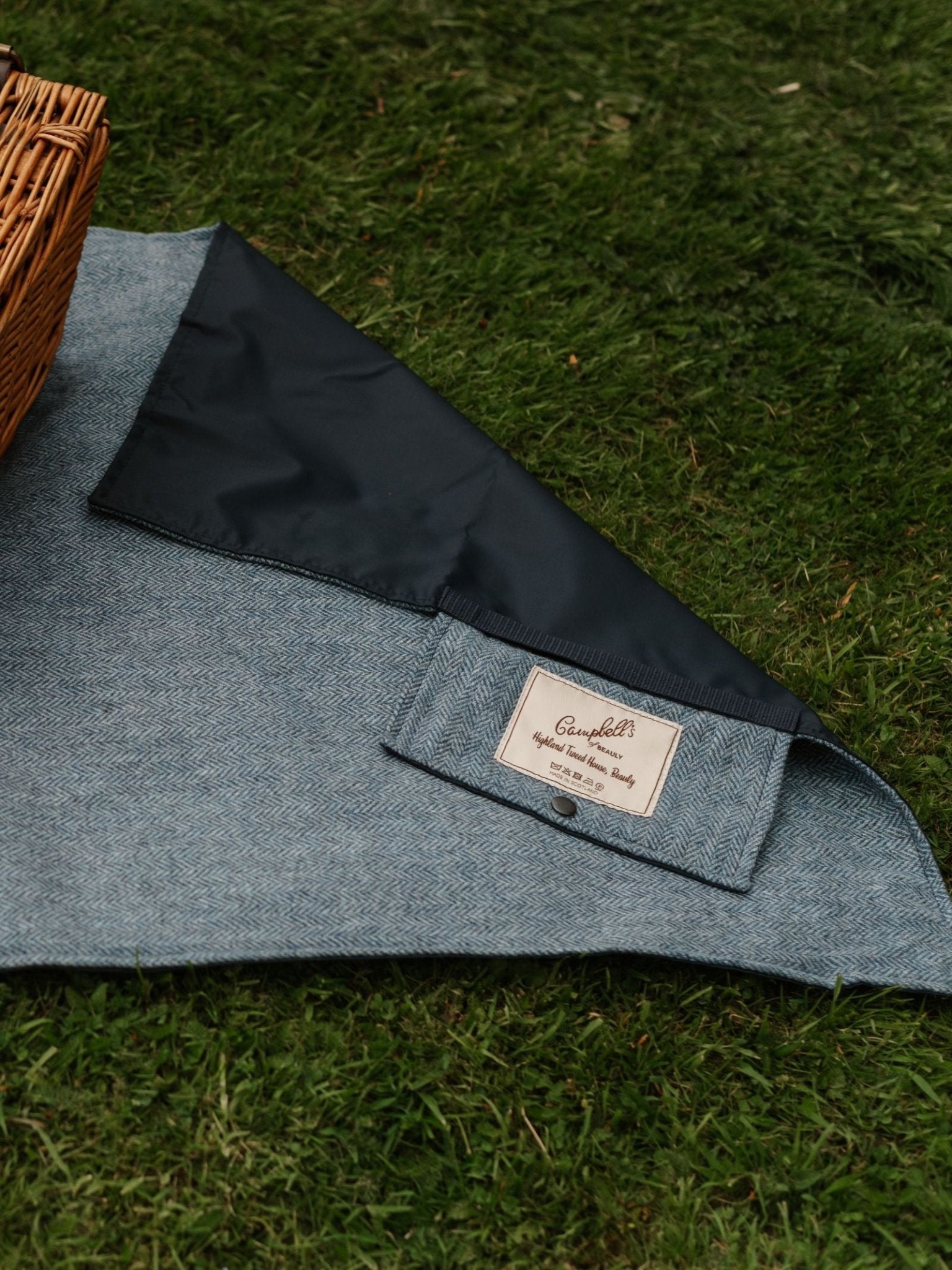 A close-up of the Walker Camper Picnic Rug by Campbell's of Beauly, featuring gray fabric and black waterproof lining, partially folded on green grass beside a wicker basket. The Campbell's of Beauly label is visible on this stylish picnic rug.