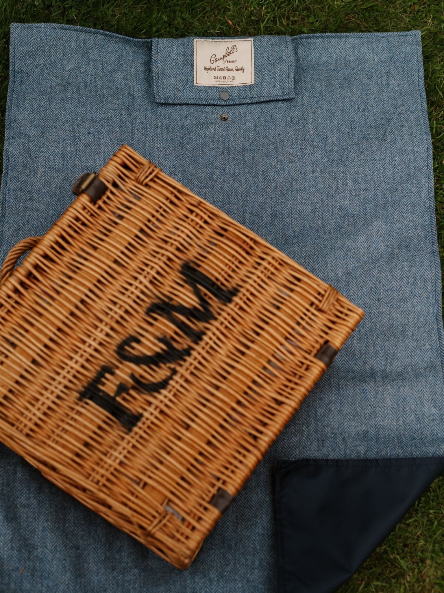 A wicker picnic basket labeled F&M rests on a Campbell's of Beauly Walker Camper Picnic Rug, which is blue patterned and spread out on green grass.