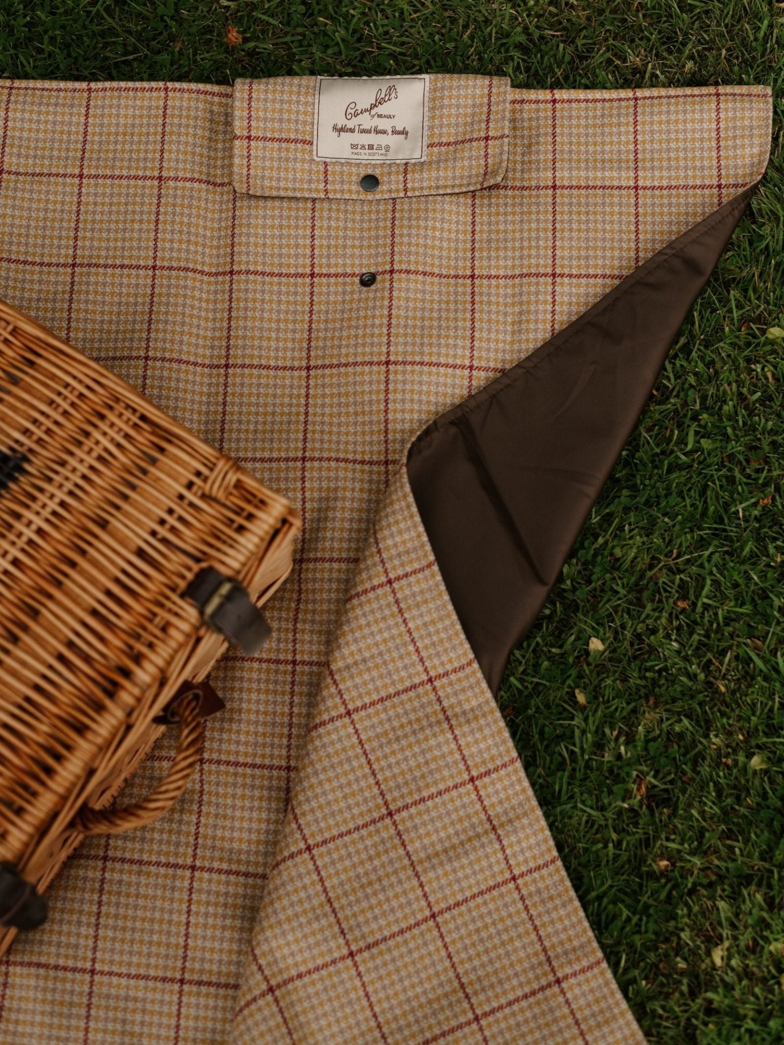 A close-up of the Campbell's of Beauly Walker Camper Picnic Rug, featuring a beige and red check pattern, dark brown underside, and label, partially folded on grass beside a wicker basket—perfect for countryside style.
