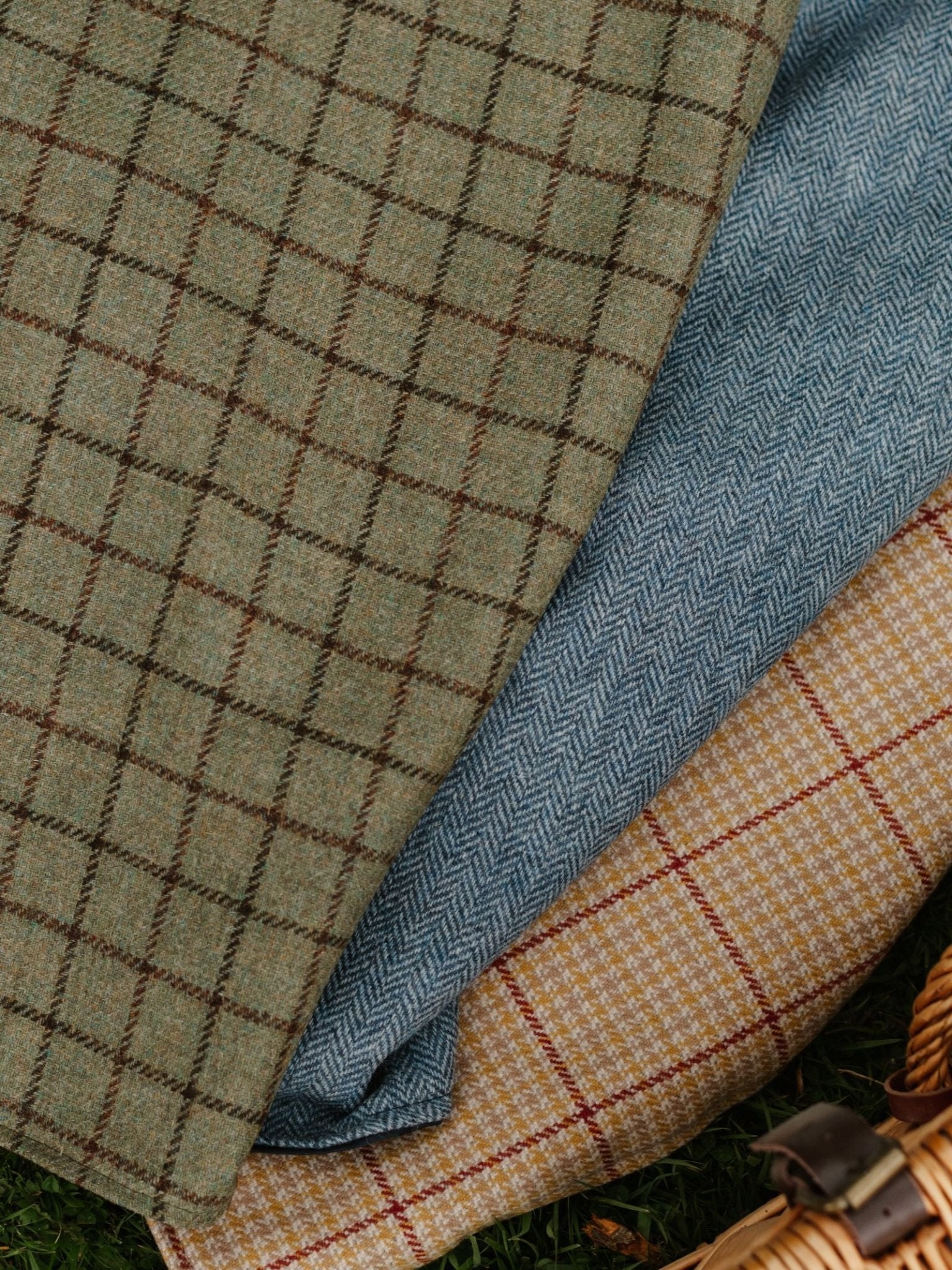 A close-up of three overlapping Walker Camper Picnic Rugs by Campbell's of Beauly in Brown 611, each with a unique pattern and waterproof backing. A picnic basket is partially visible in the lower right corner.