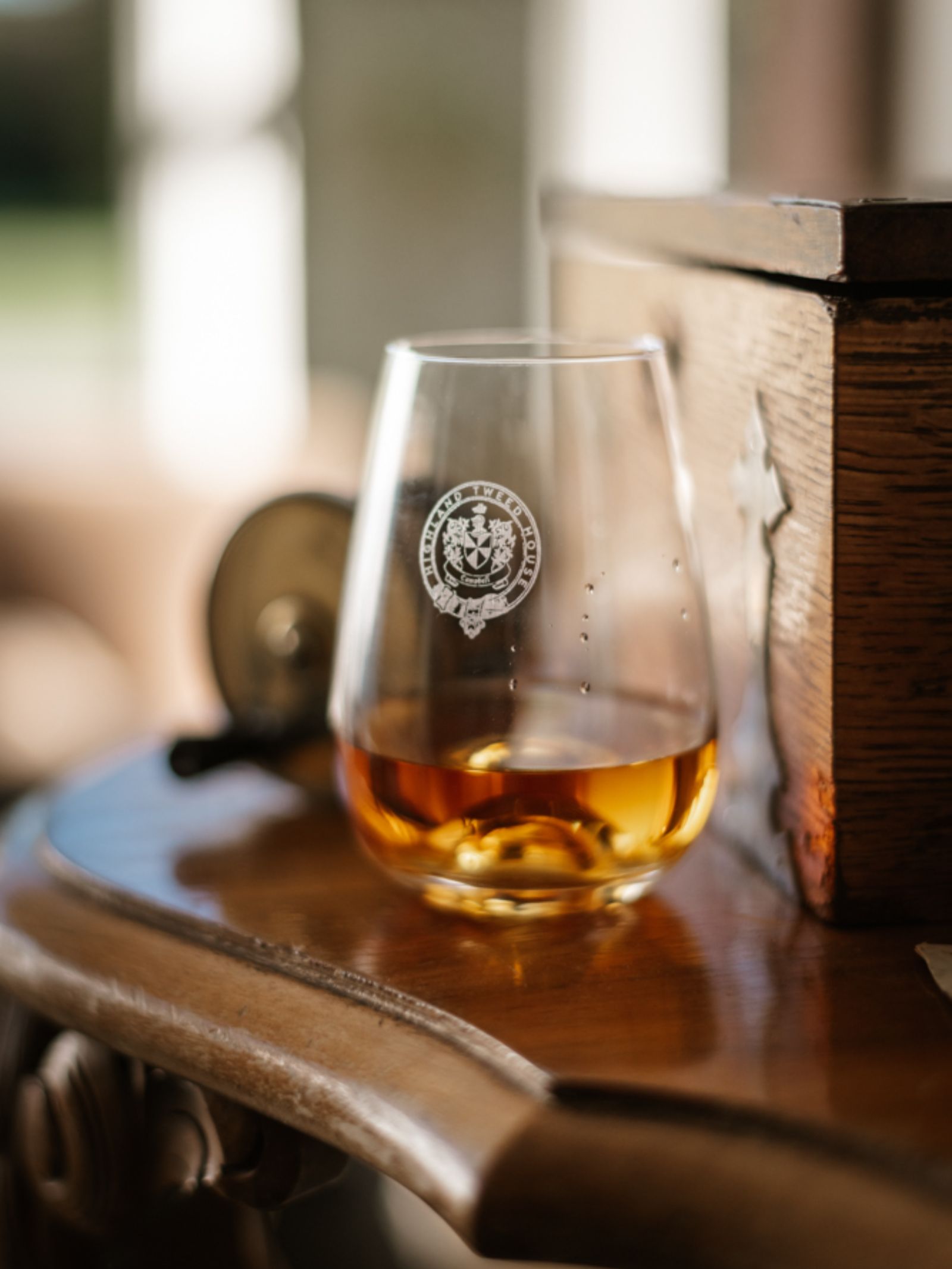 The Campbell's of Beauly Whisky Tasting Glass, with its tulip shape and etched emblem, rests beside a wooden box on a polished table in sunlight—ideal for whisky fans seeking a refined tasting experience.