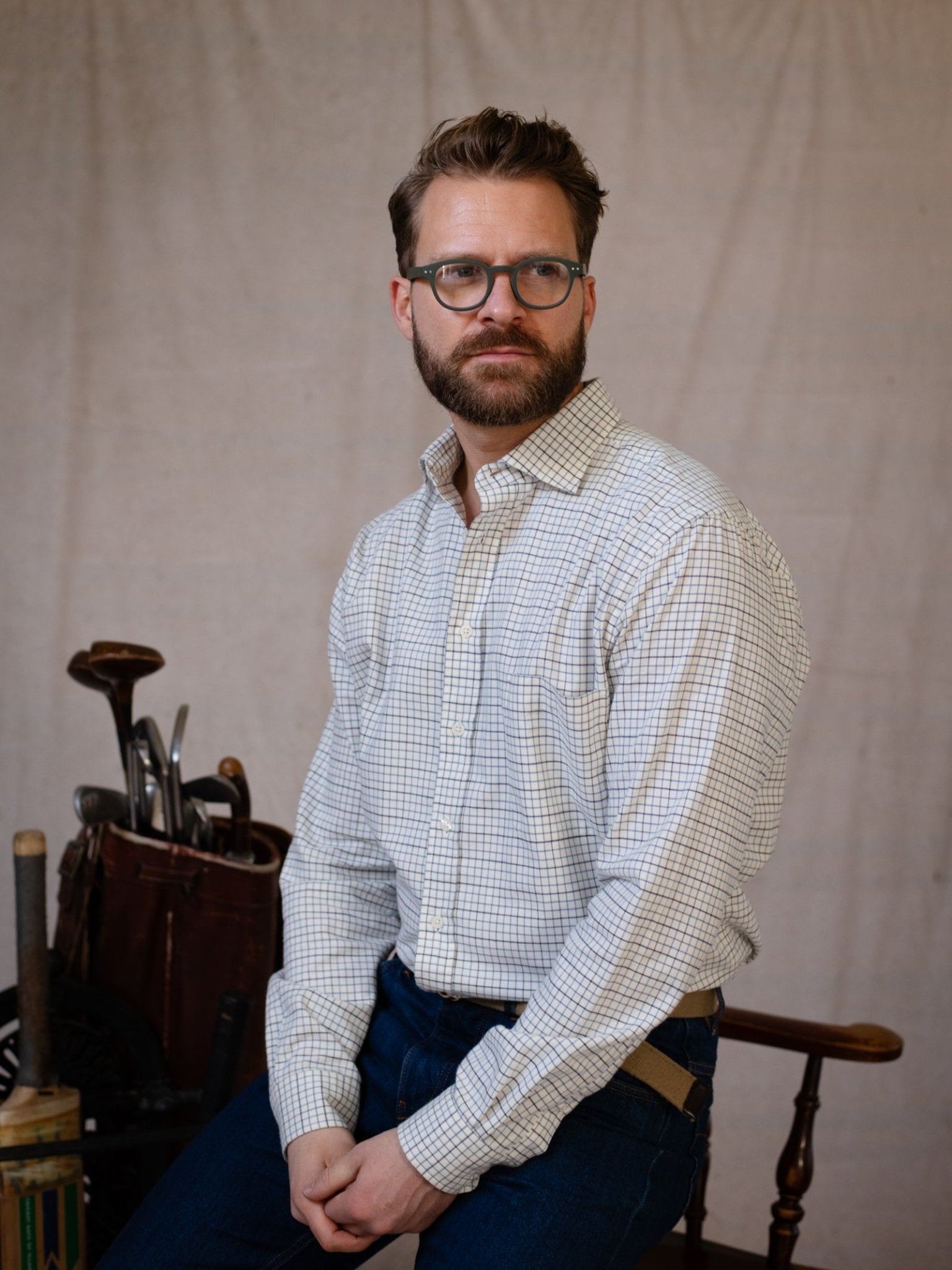 A man with glasses and a beard wears the Campbell's of Beauly Windowpane Shirt and blue jeans while seated on a wooden chair; golf clubs in a bag are visible in the background against a plain backdrop.