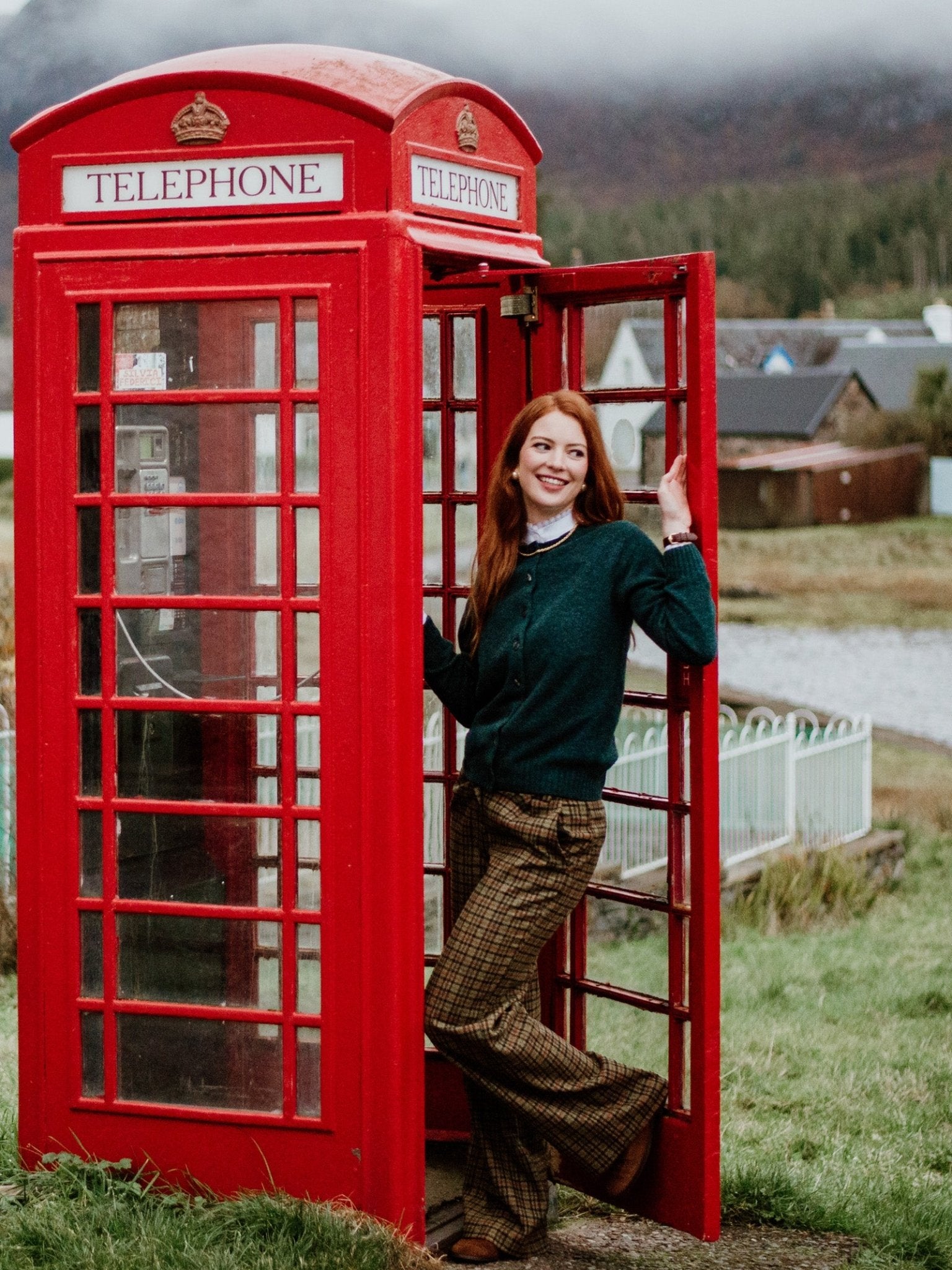 A smiling woman with long red hair stands in a classic red British phone booth, wearing the Campbell's of Beauly Auld Stock Geelong Lambswool Crew Cardigan, with grassy fields and misty hills in the background.