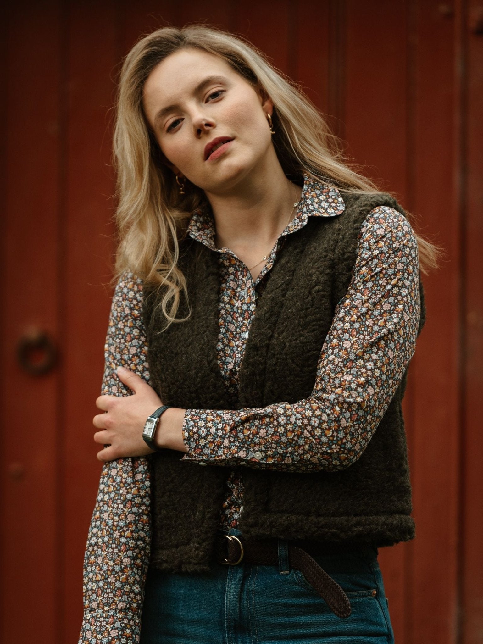 A woman with long blonde hair stands before a red door, wearing Campbell's of Beauly's Wool Shrug over blue jeans and a floral shirt, paired with a silver watch—showcasing lightweight warmth ideal for any season.
