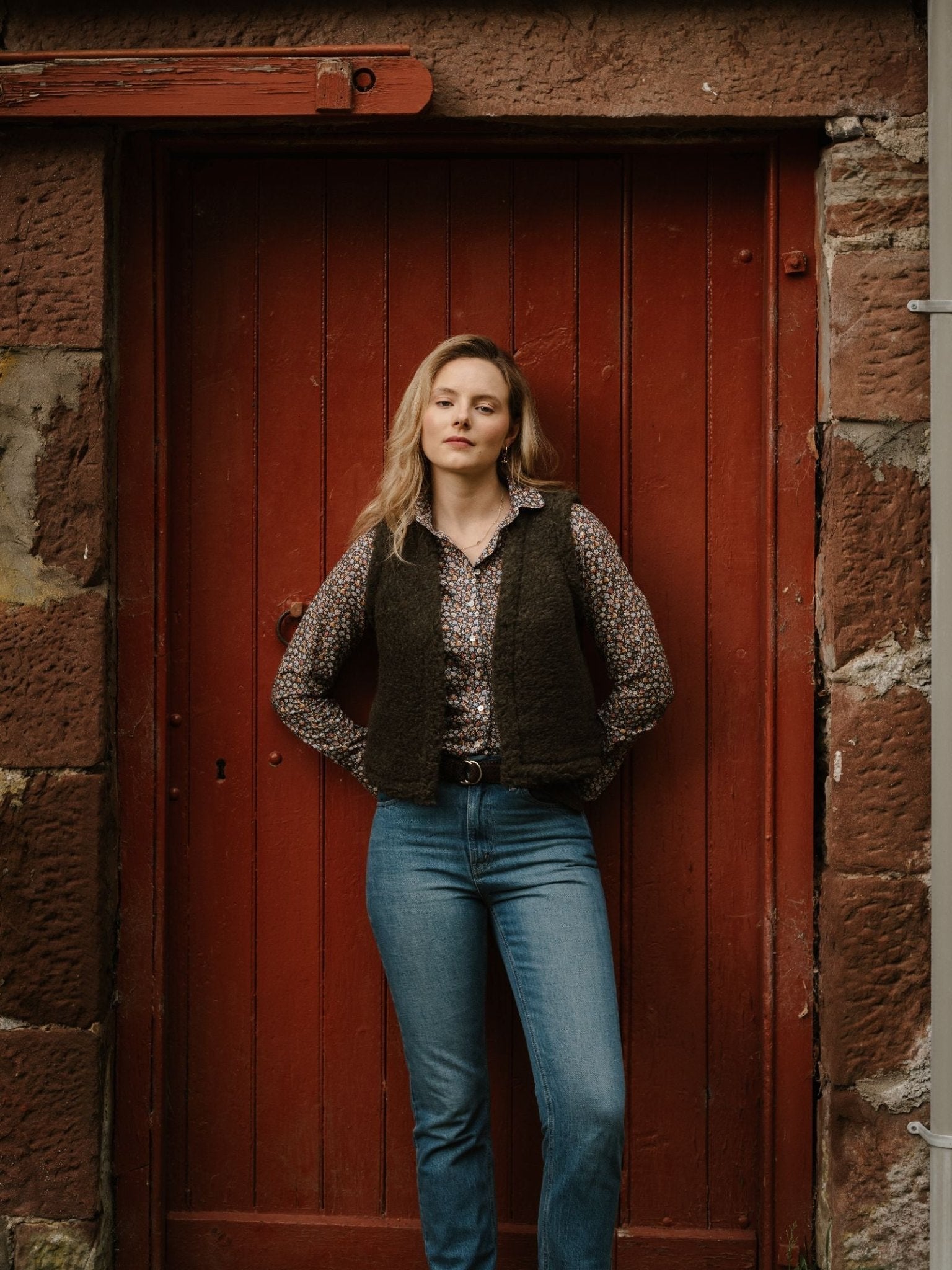 A woman with blonde hair showcases stylish layering in a Campbell's of Beauly Wool Shrug, floral shirt, and blue jeans as she stands hands-in-pockets before a rustic red wooden door framed by stone walls.