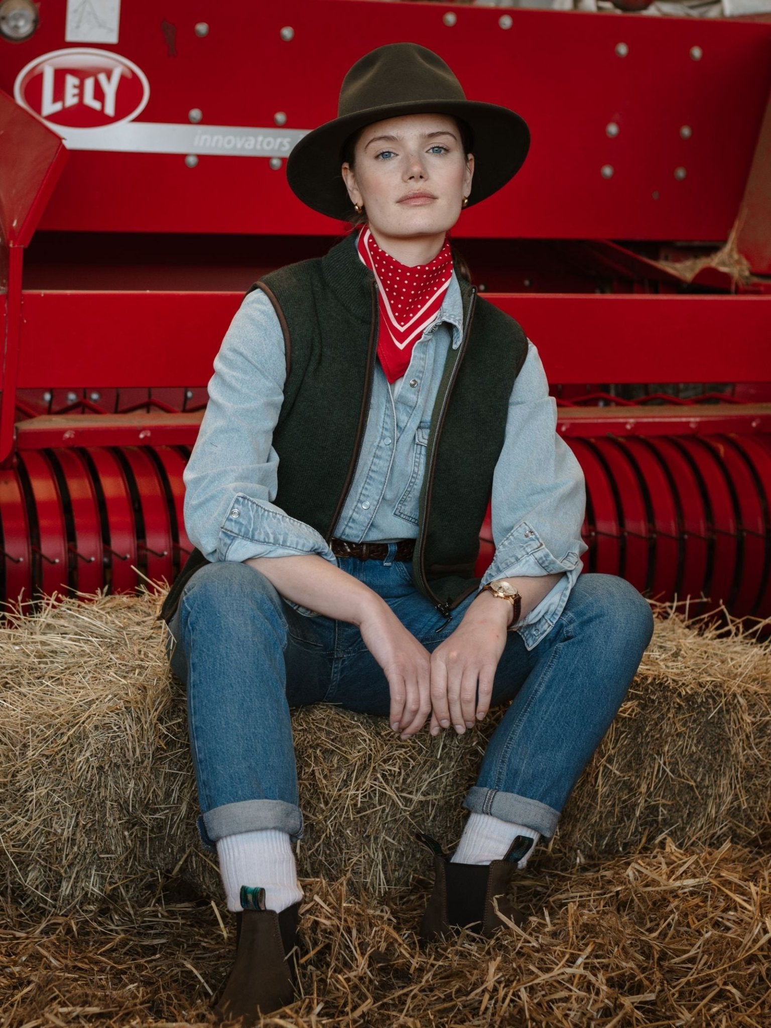 A person in a wide-brimmed hat, red bandana, denim shirt, Campbell’s of Beauly Merino Cashmere Gilet, jeans, and boots sits on a hay bale by red farm equipment, showcasing countryside style and confidence.