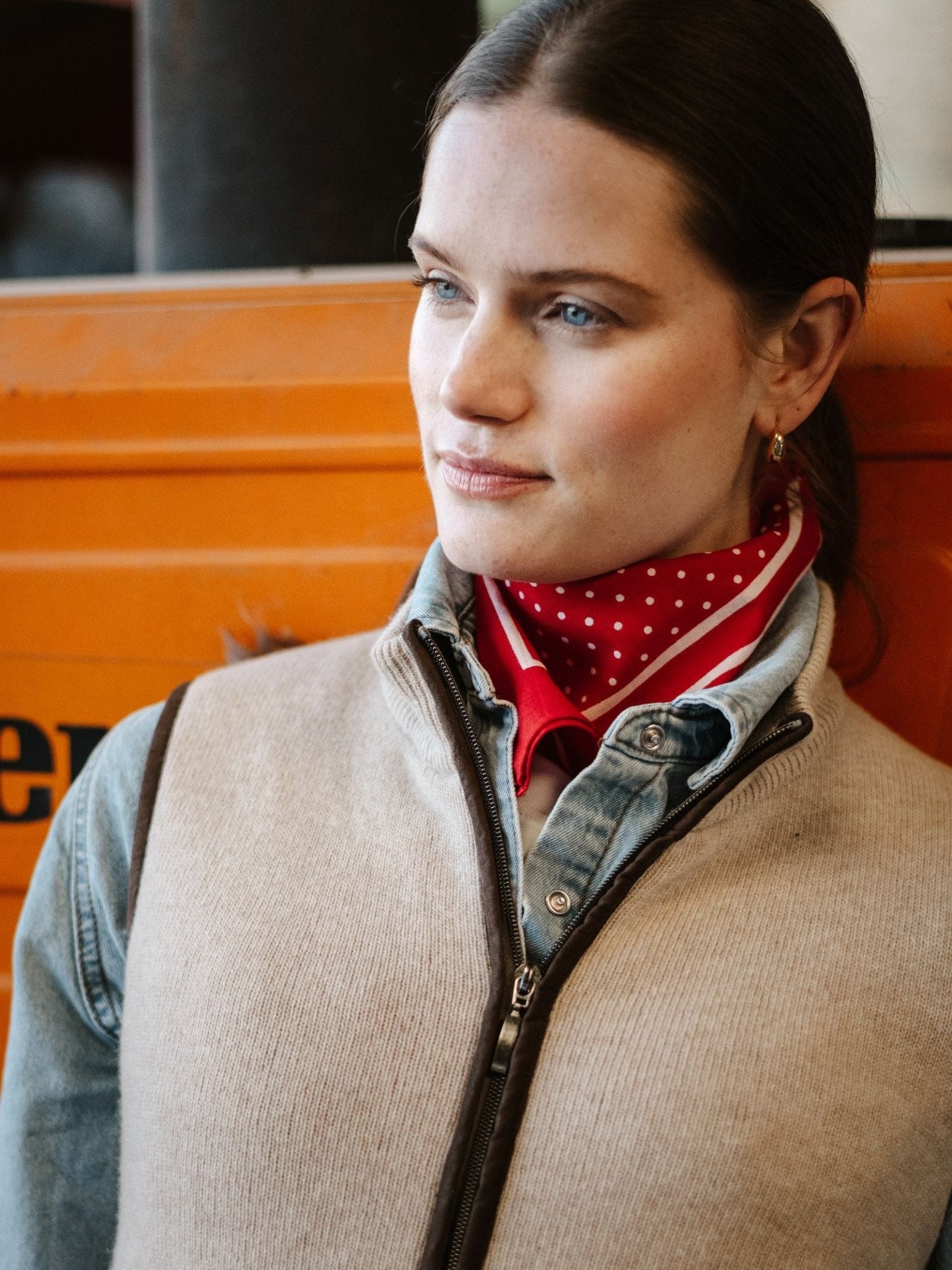 A woman with light skin and blue eyes wears the Campbell's of Beauly Merino Cashmere Gilet over a denim shirt and a red polka dot scarf. She stands calmly before an orange background.